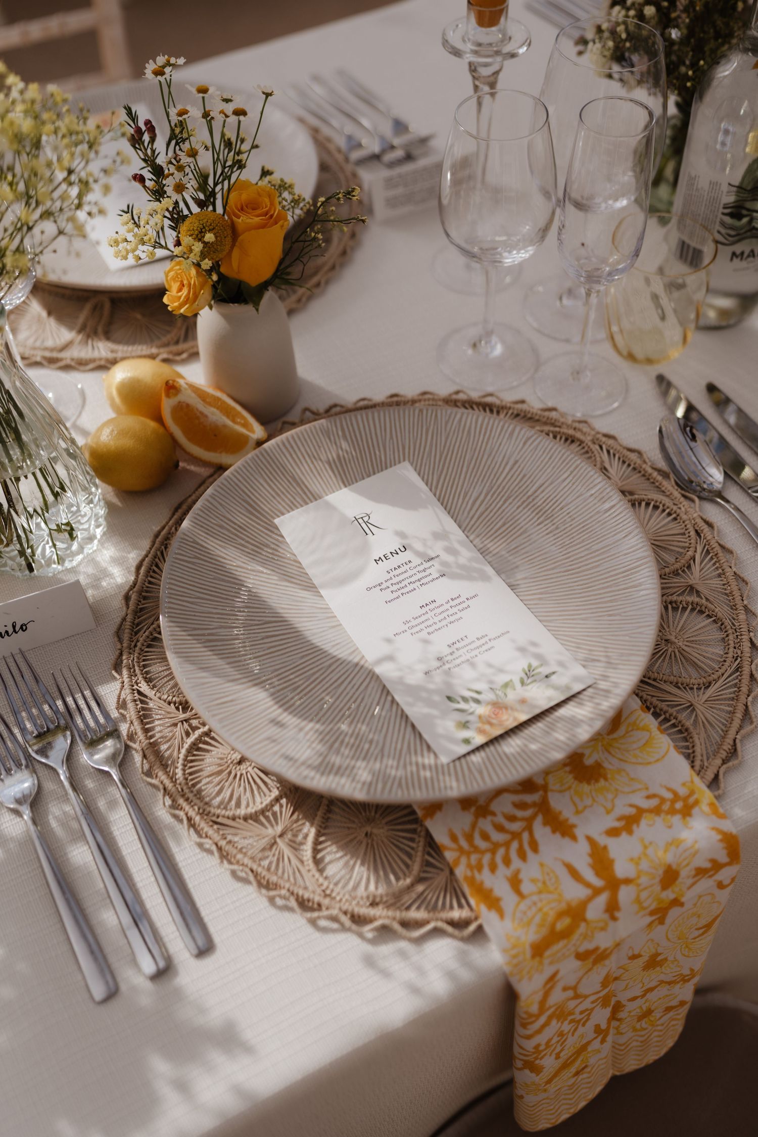 Elegant wedding table setting with rattan charger, menu card, yellow flowers, lemons, and floral napkin.