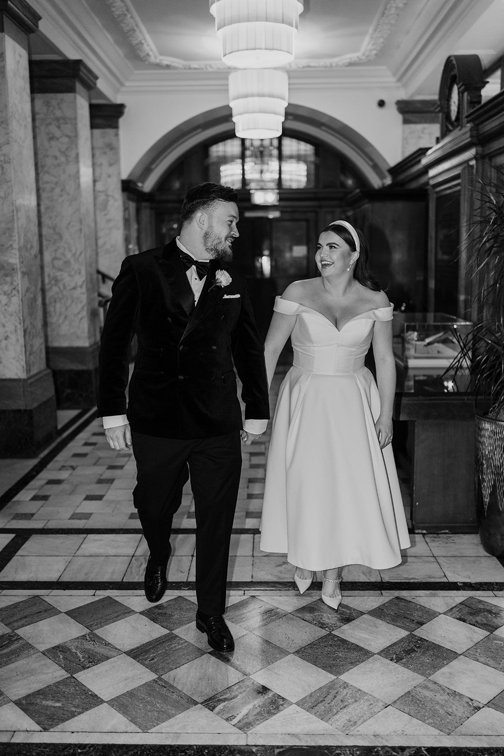 Black and white photo of a couple walking in a grand hallway with checkered floors and elegant chandeliers.