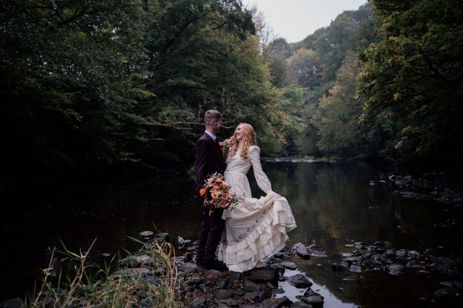 Couple dancing on their wedding day stood on rocks in a river with trees surrounding them.
