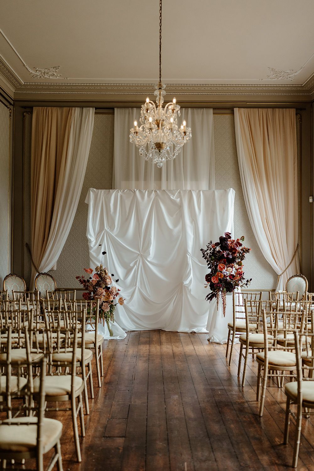 Autumnal wedding flowers with red, burgundy, pink and white flowers styled into a ceremony space with draping in Rise Hall.