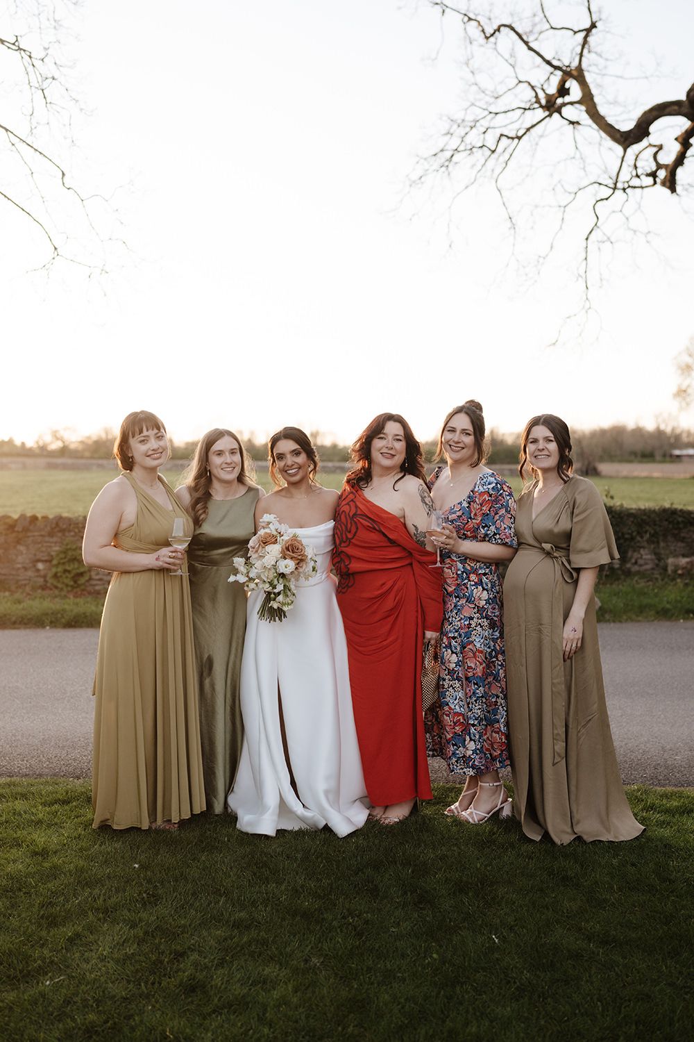 Elegant bridal party poses outdoors at sunset wearing mix of olive green, red and floral dresses on a grassy lawn.