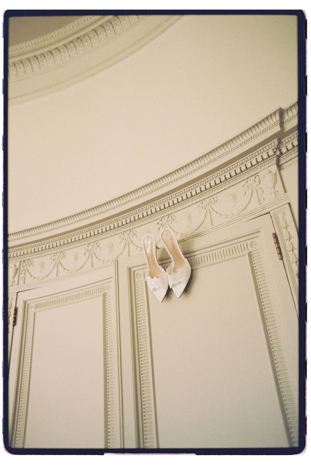White bridal heels hanging on ornate cream wall paneling with decorative molding in an elegant wedding venue.