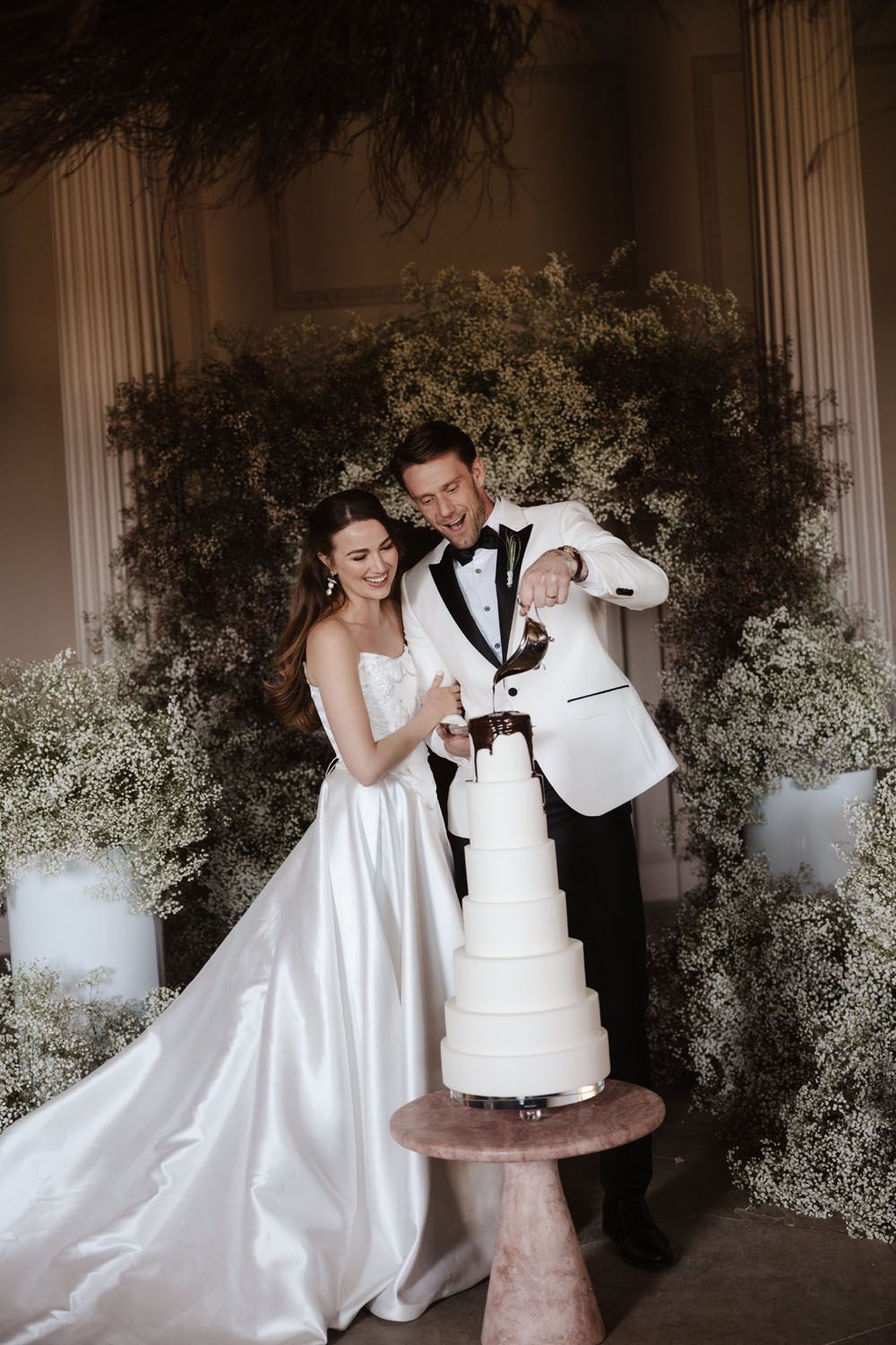 Bride and groom cutting a tall white wedding cake surrounded by lush greenery and baby's breath floral backdrop.