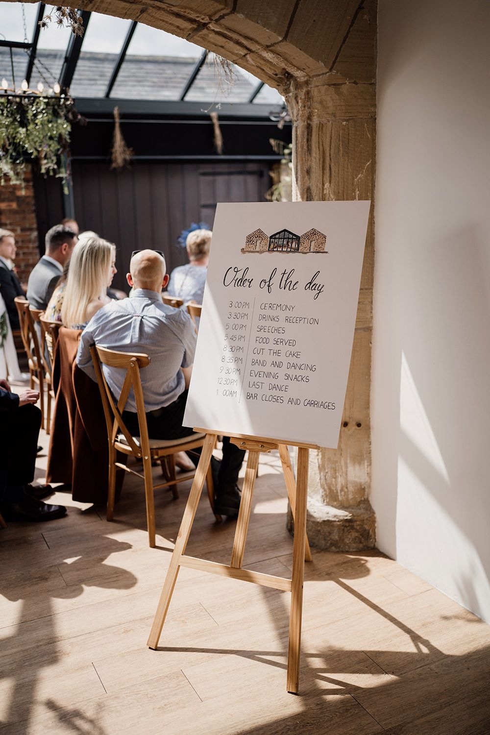 Illustration of the wedding order of the day on a wooden easel with guests sat behind in the ceremony room.