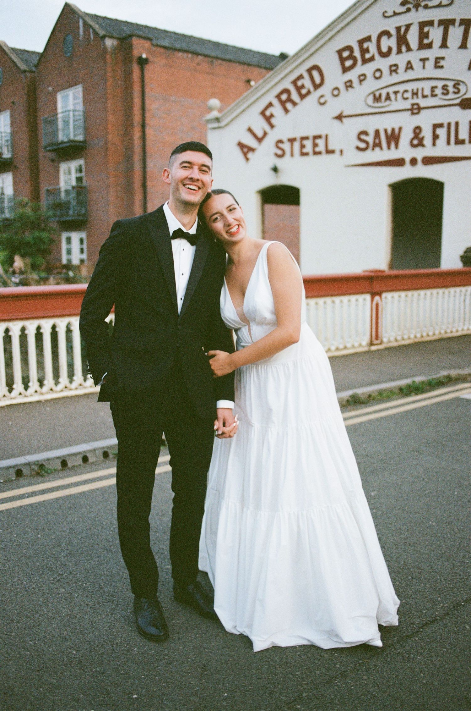 A couple in wedding attire poses in front of a vintage industrial building with 'Alfred Beckett' steel works signage.
