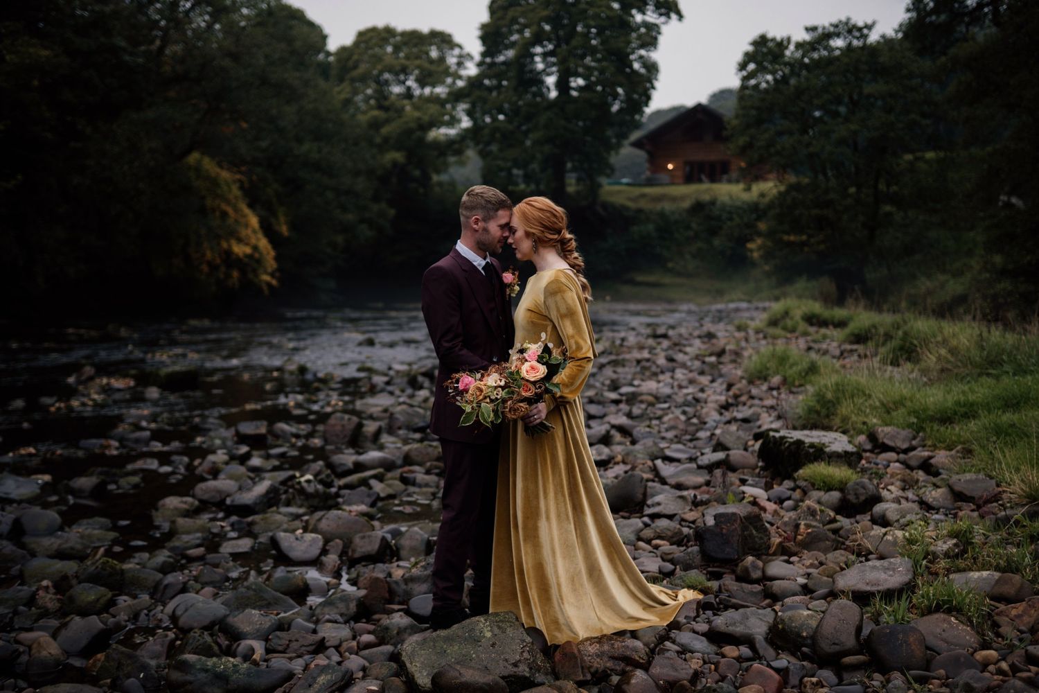 Couple kissing in autumnal styling while stood on rocks at Hidden River Barn with trees surrounding them.
