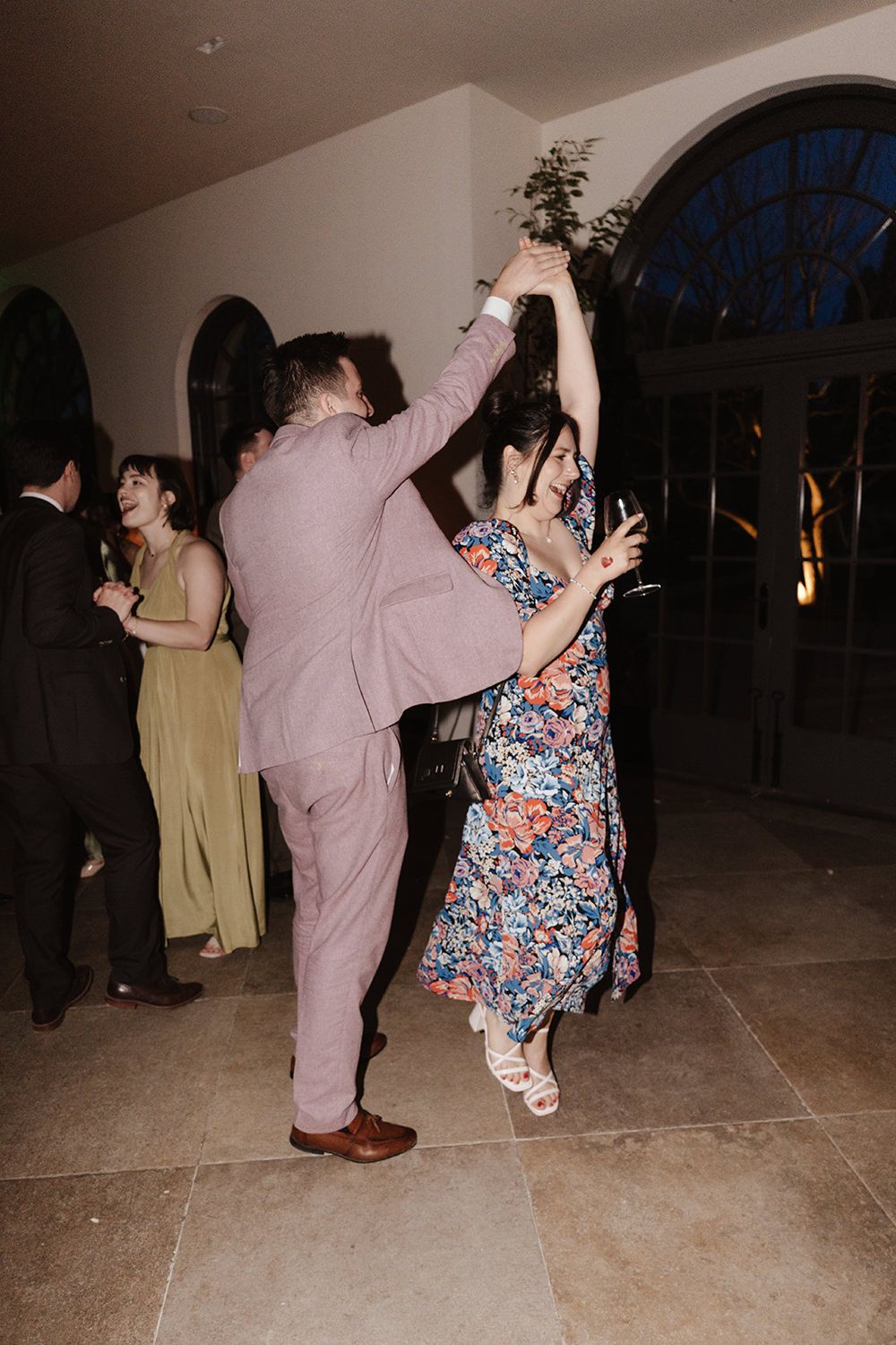 Dancers at a formal event swing dancing energetically on a tile floor with arched windows in the background.