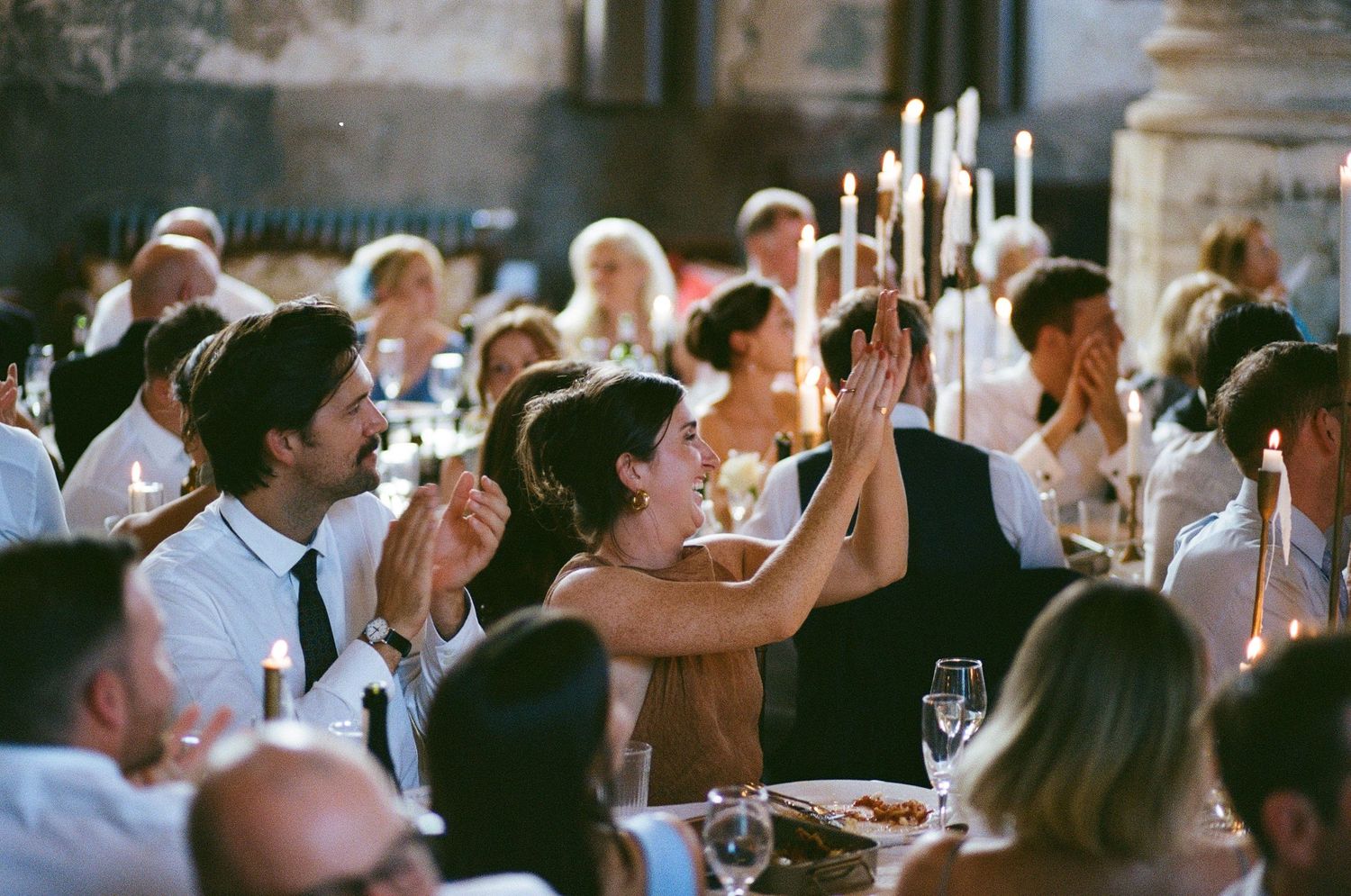 Wedding guests clap and celebrate joyfully at a candlelit reception dinner table.