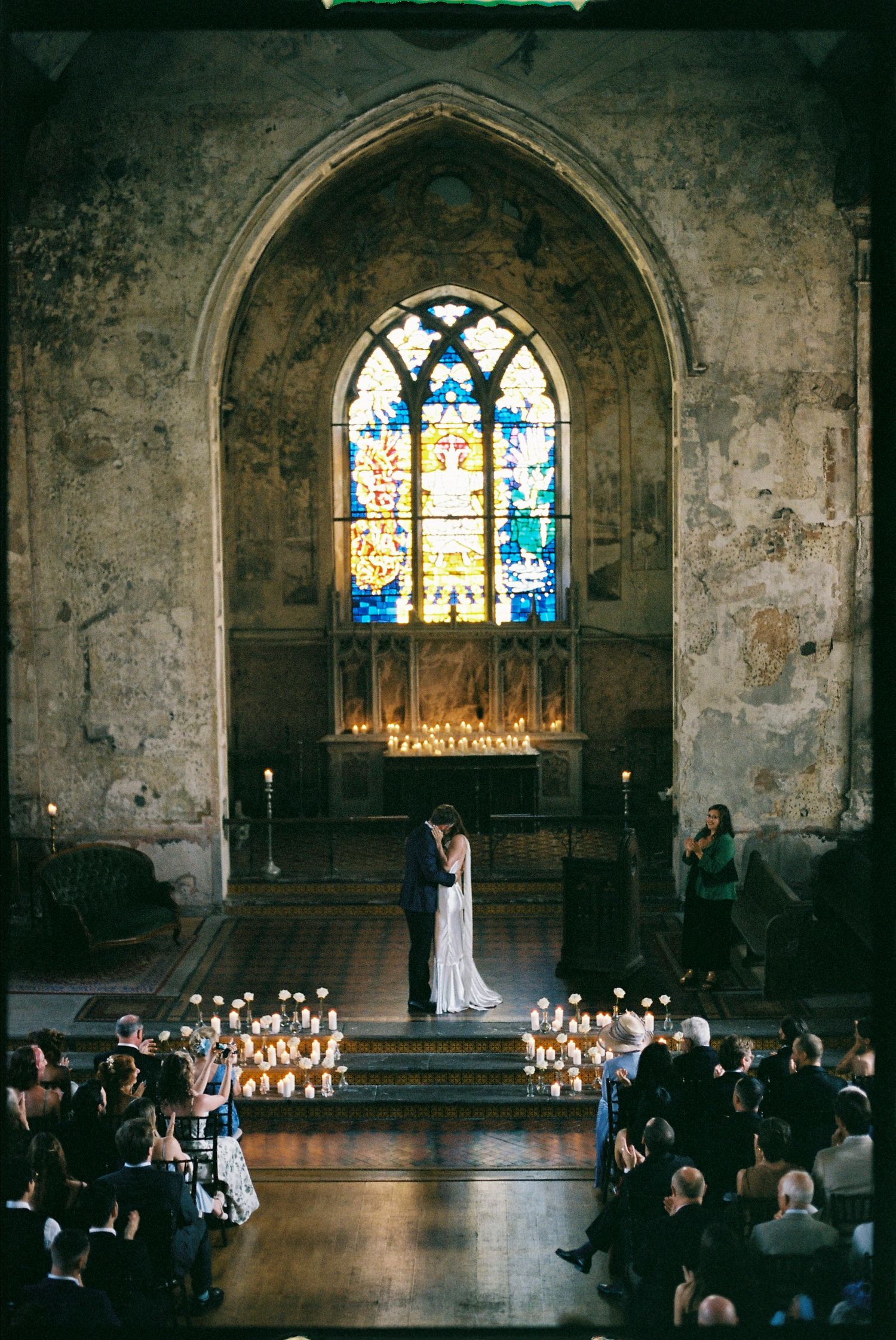 Bride and groom share a kiss at a candlelit church altar beneath a stained glass window.