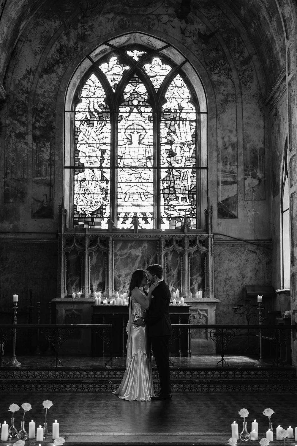 A silhouetted couple embraces in a church beneath a dramatic gothic stained glass window in black and white.