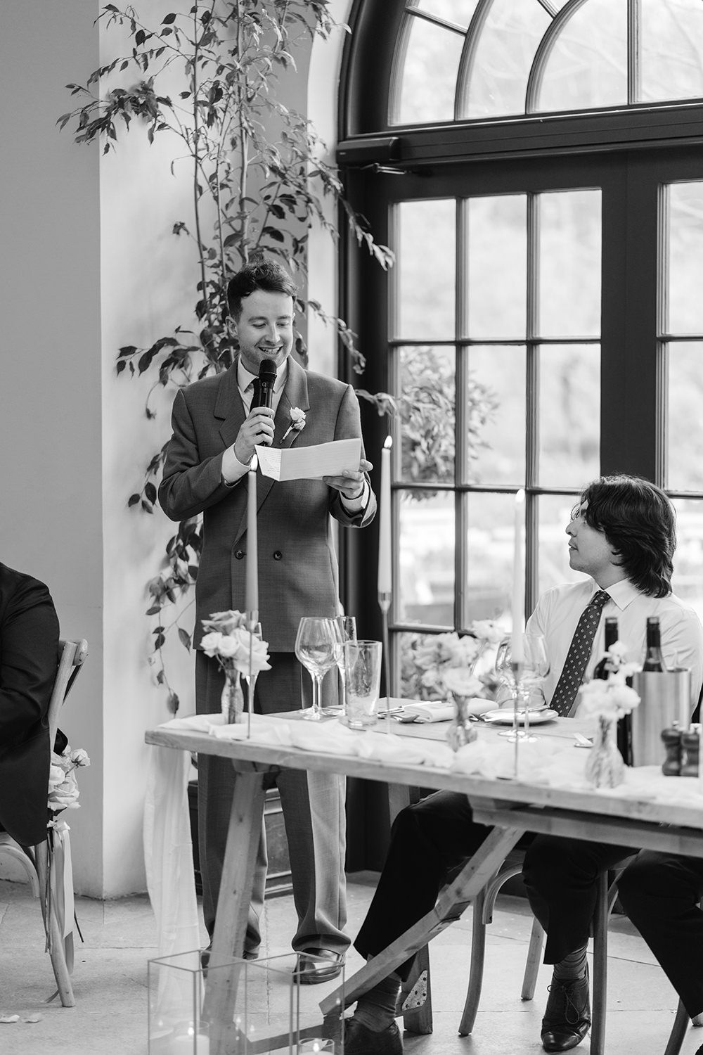 A wedding celebration showing speech-giving during a reception in a room with arched windows and greenery.
