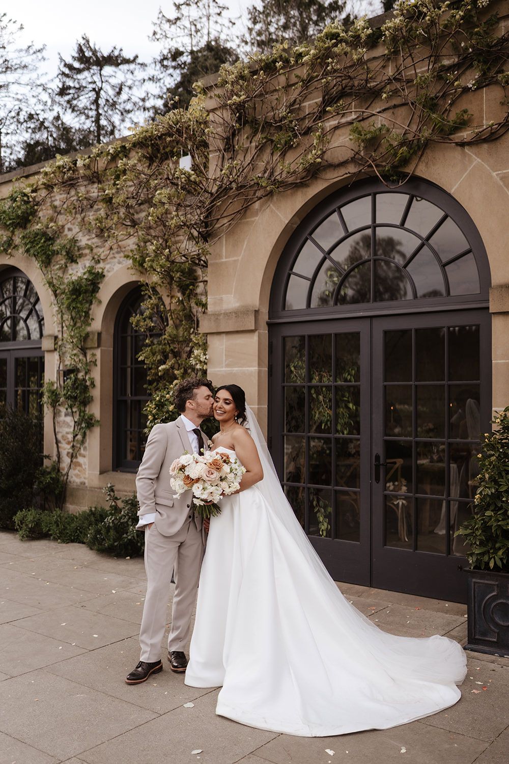 A wedding couple shares a romantic moment in front of elegant arched doors with climbing vines at a stone building.