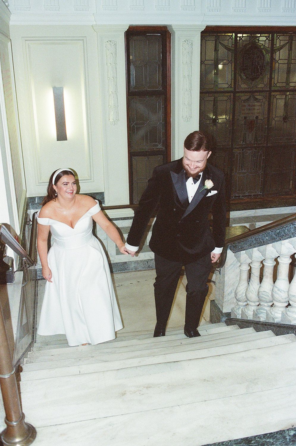 A bride in a strapless white gown and groom in a black tuxedo descend an elegant white staircase together.