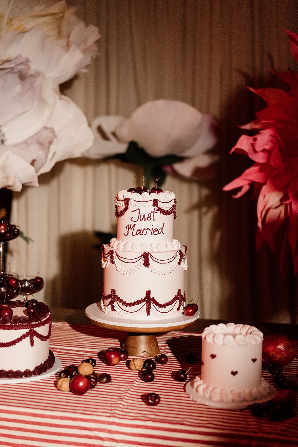 White wedding cakes with red ribbon decorations displayed on a striped tablecloth at a reception dessert table.