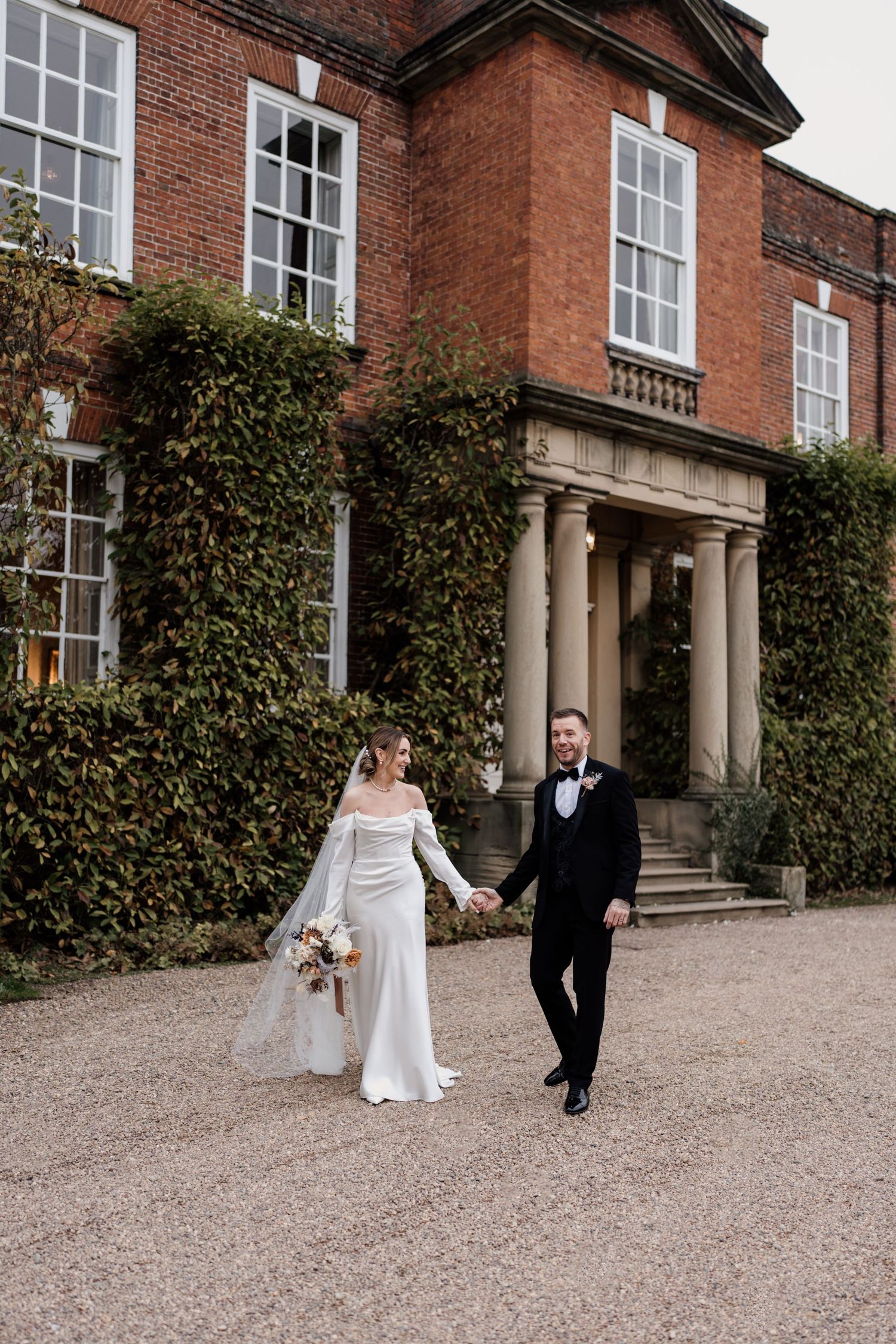 A couple walks on gravel outside an elegant red brick Georgian manor house with ivy-covered walls and white columns.