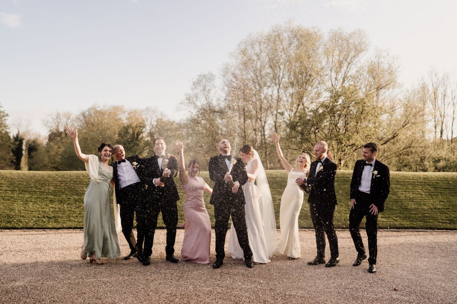 Wedding party poses joyfully on path with autumn trees in background during golden hour.