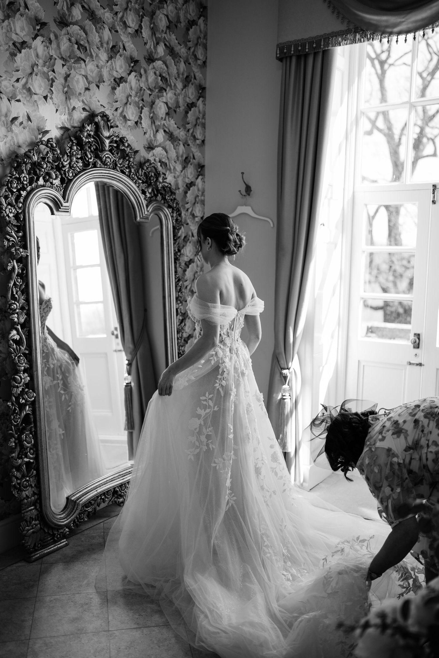 Bride getting into her wedding dress looking into a mirror while her mam is adjusting her veil.