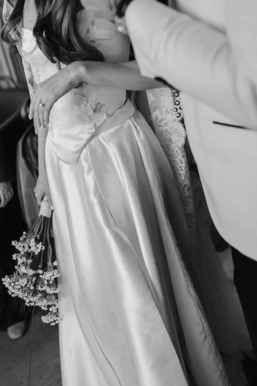 Black and white photo of groom embracing bride in satin wedding gown holding delicate floral bouquet.