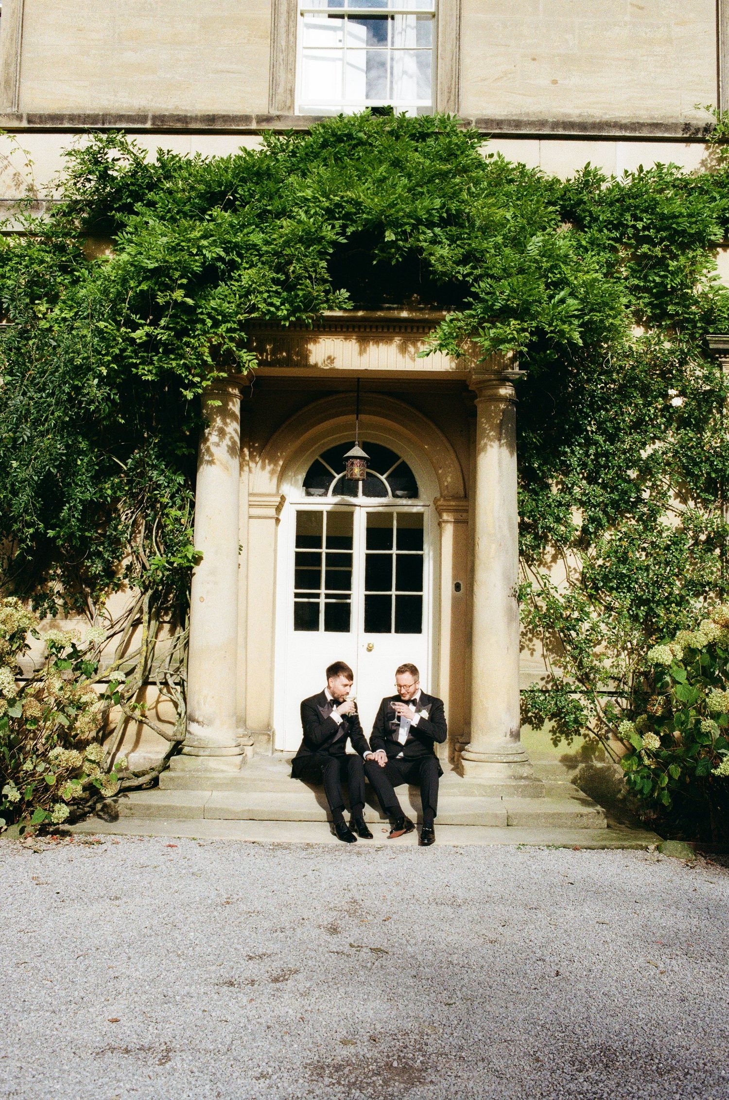 Two grooms in black suits sit together on steps of an ivy-covered stone manor house on their wedding day.