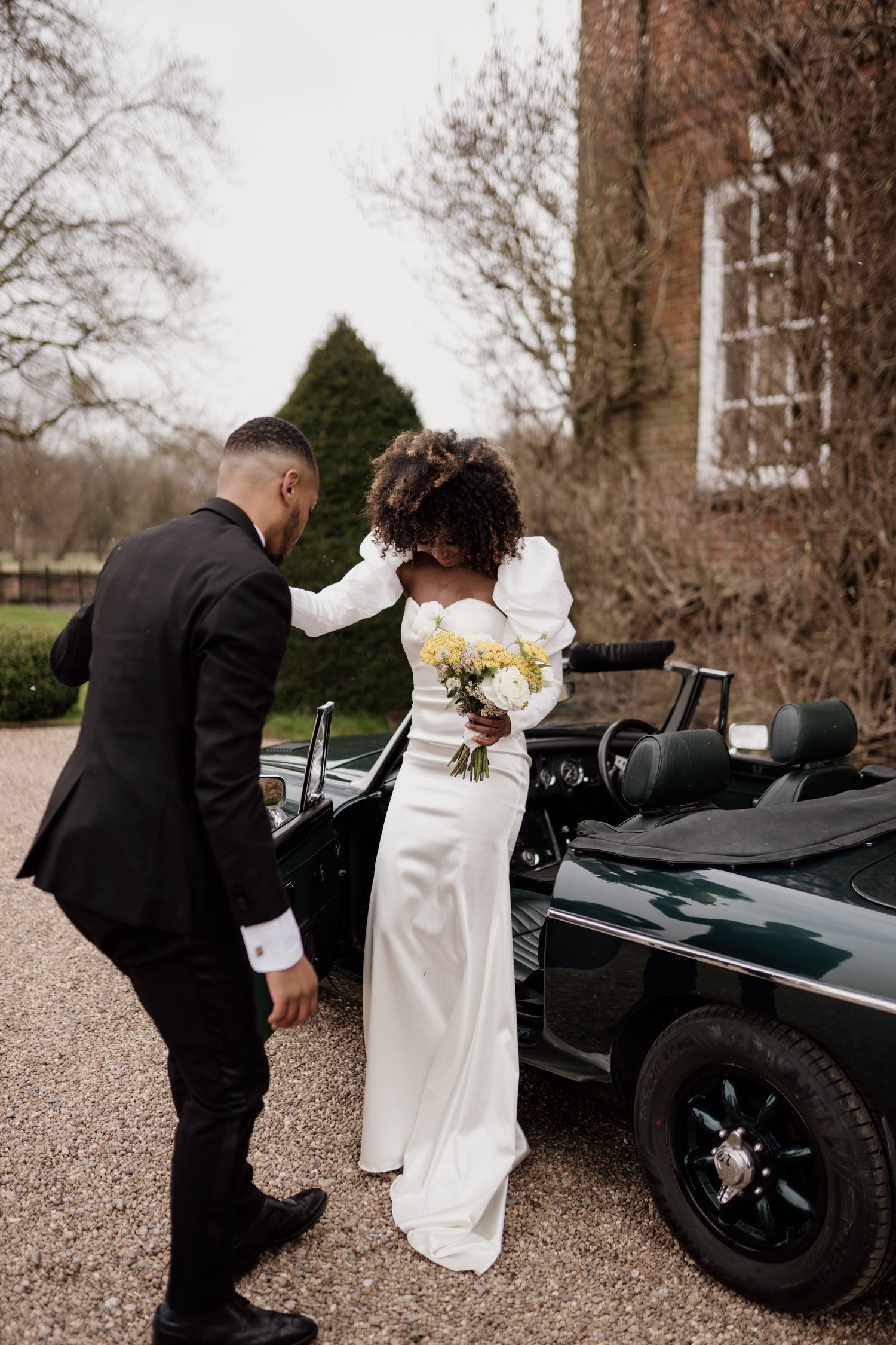 Groom assists bride in fitted white gown into a classic dark green convertible outside a brick manor.