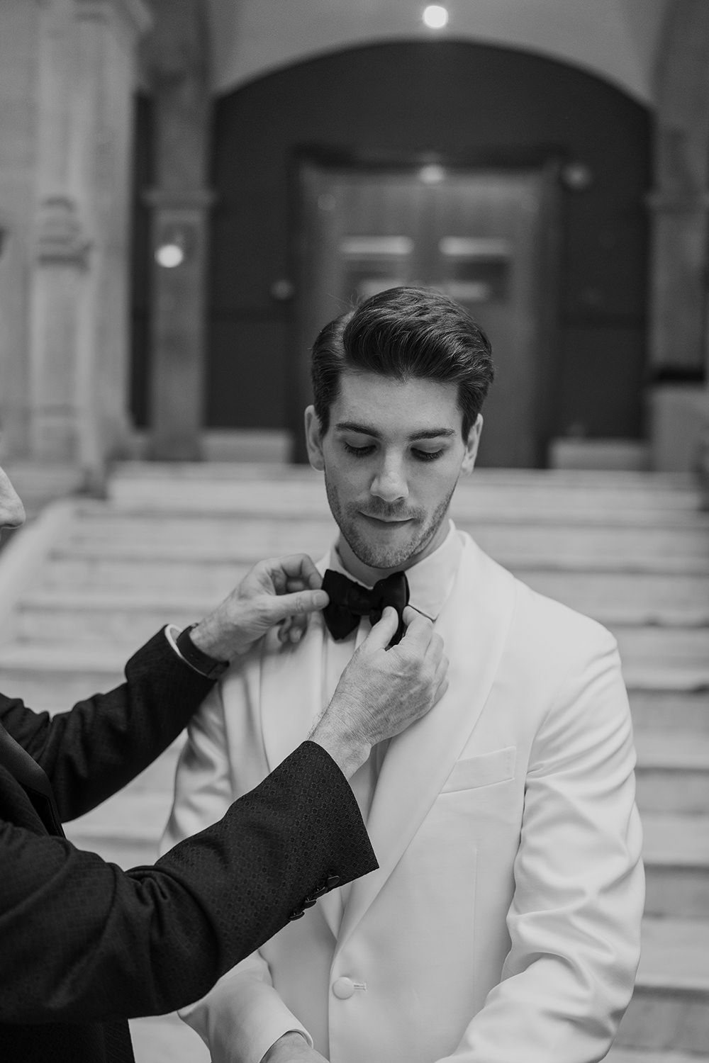 Groom looking down having his wedding tie adjusted by his dad, he's wearing a white blazer with a black bow tie.
