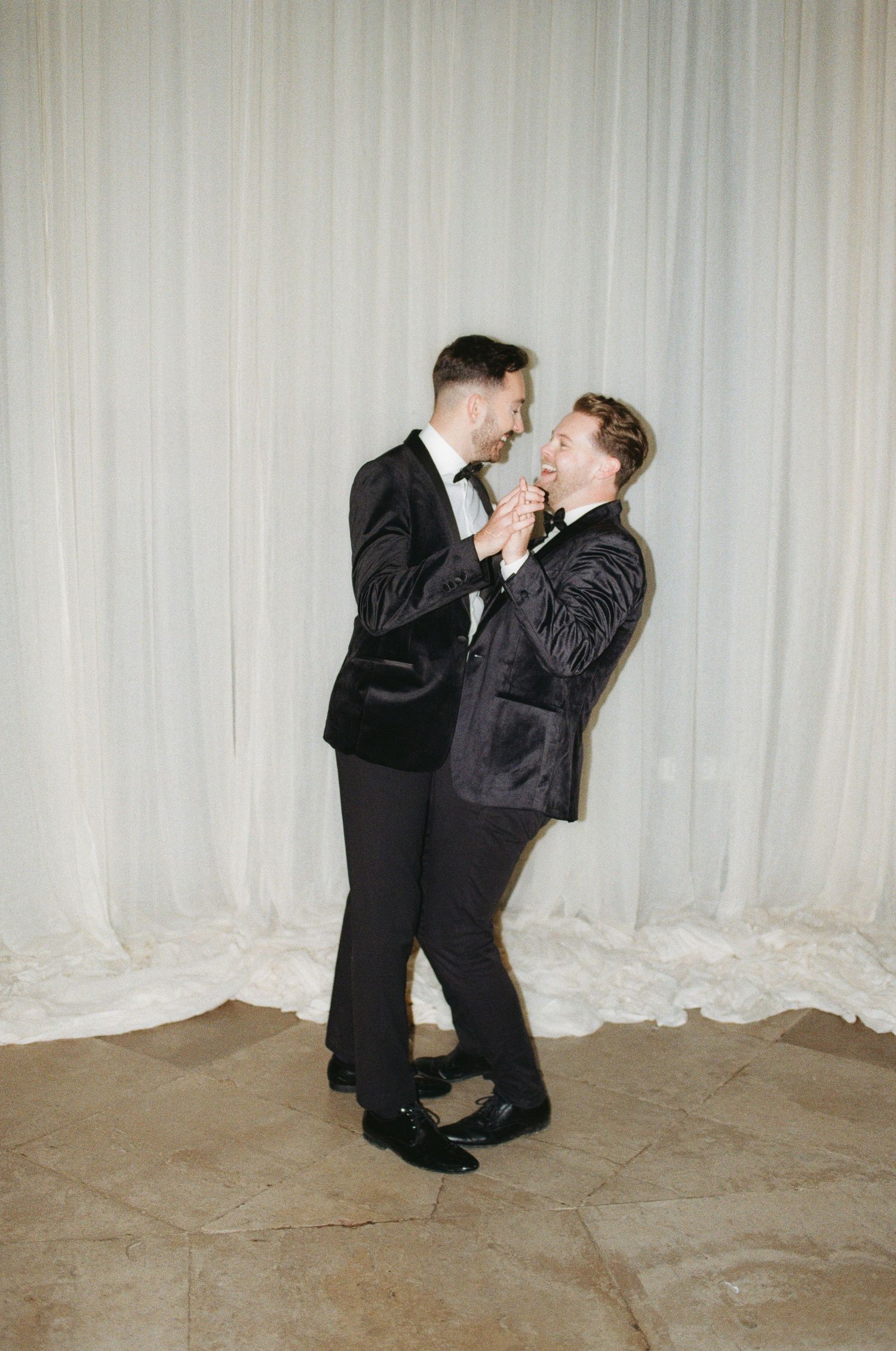 Two people in dark suits share a joyful moment dancing and laughing together against a light curtain backdrop.