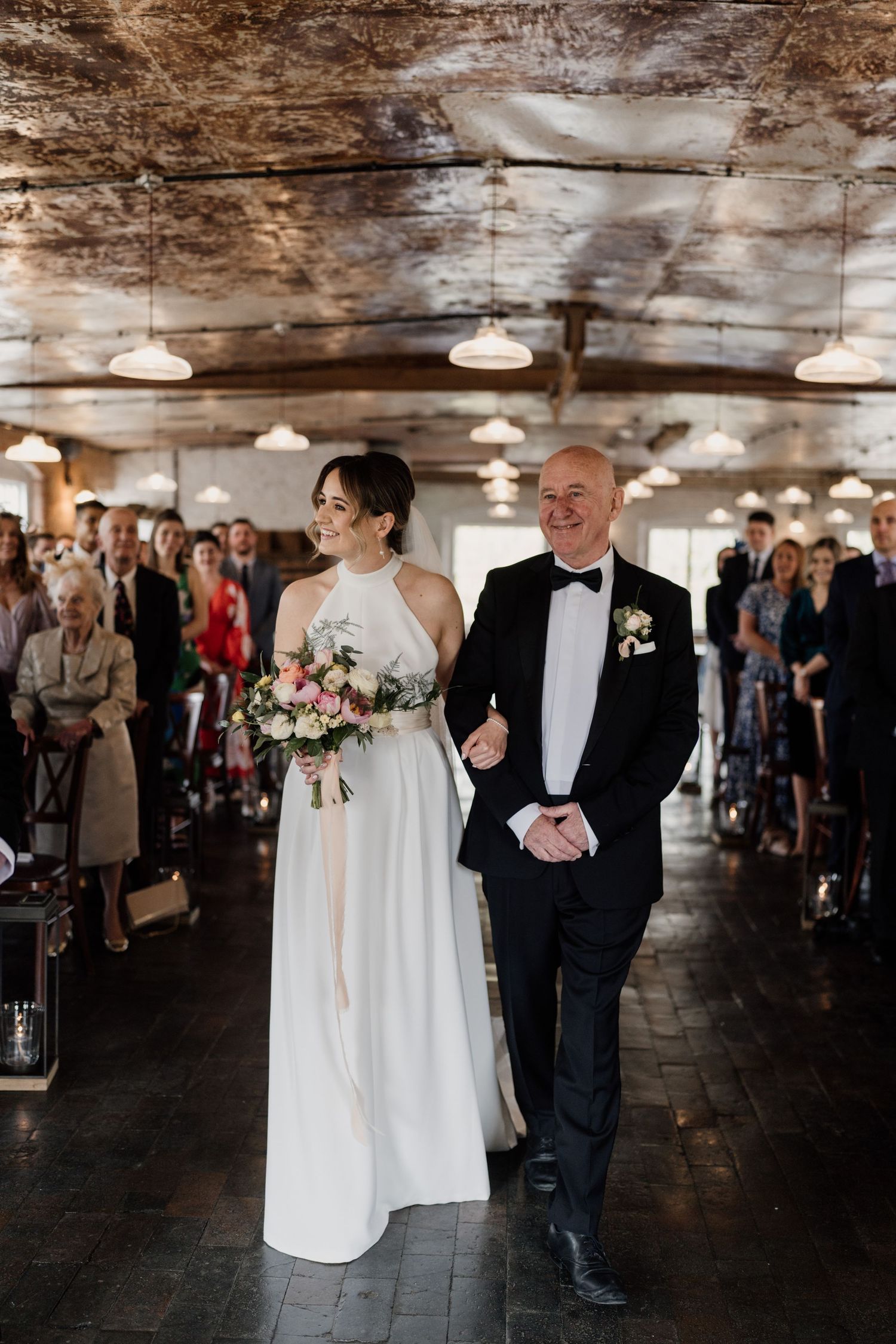 Newlyweds walking down aisle in elegant industrial venue with exposed ceiling and pendant lights.