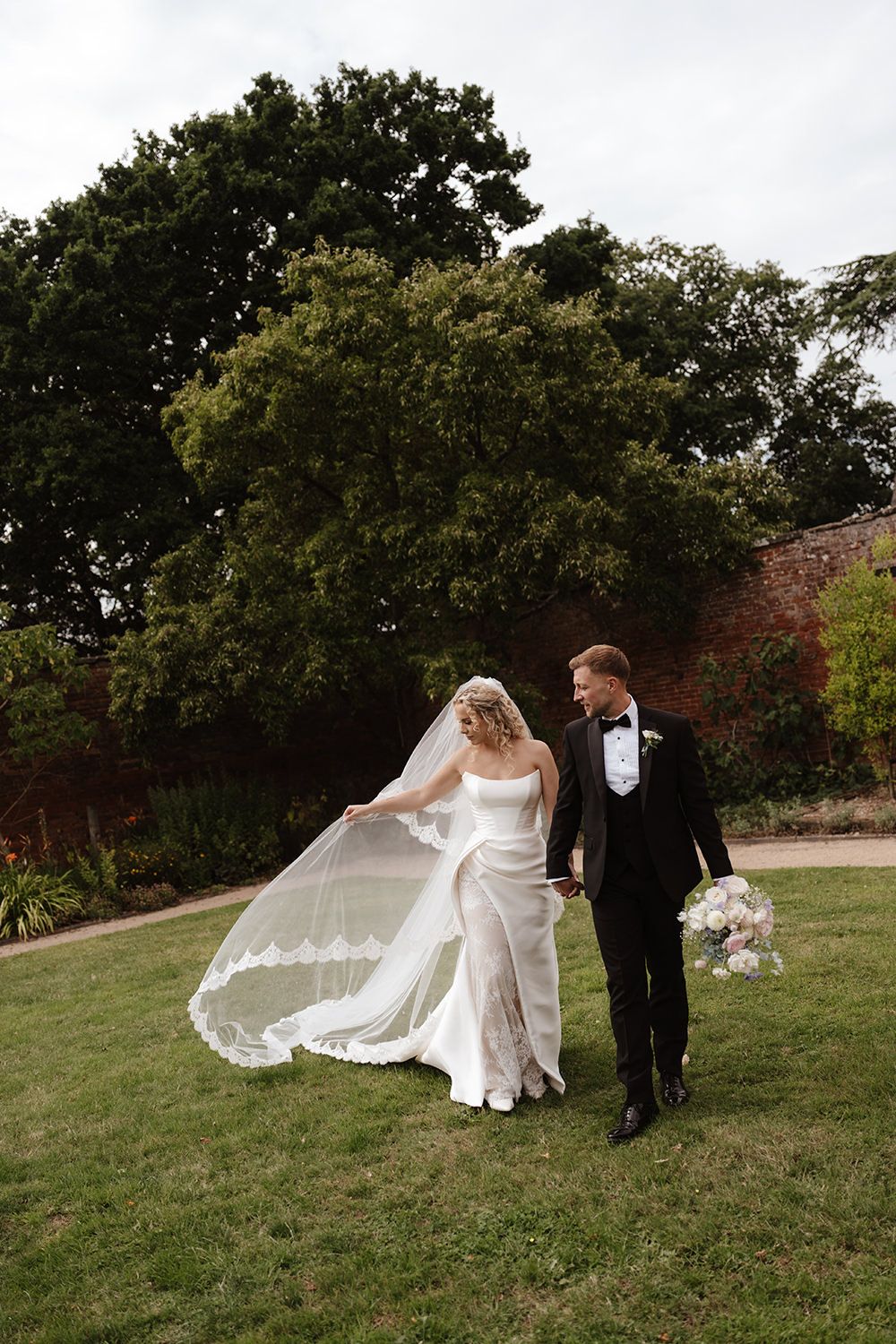 A wedding couple walks on grass with a flowing veil and lush trees in the background on their special day.