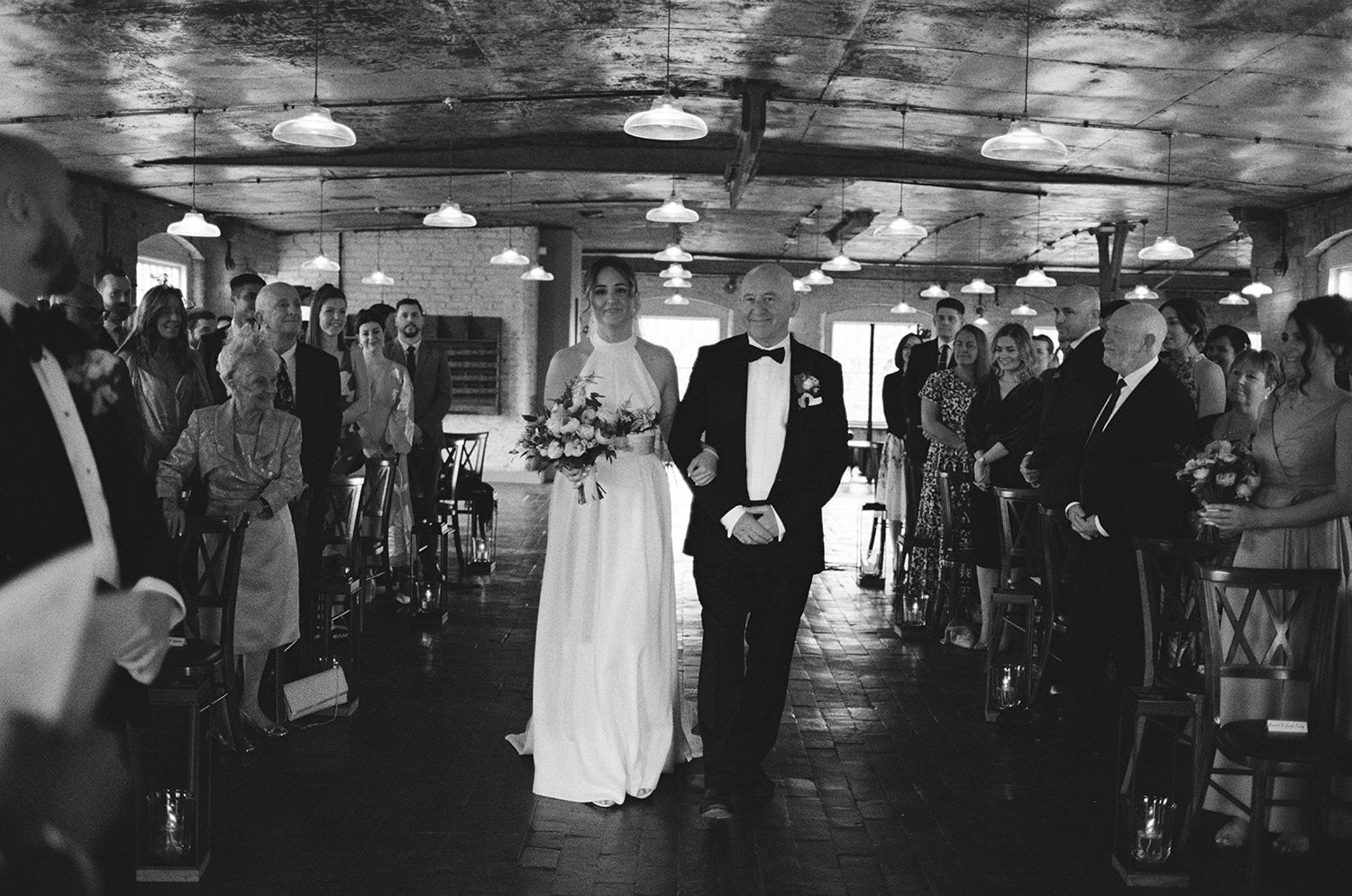 Bride holding her fathers hand as she walks down the aisle with guests watching.