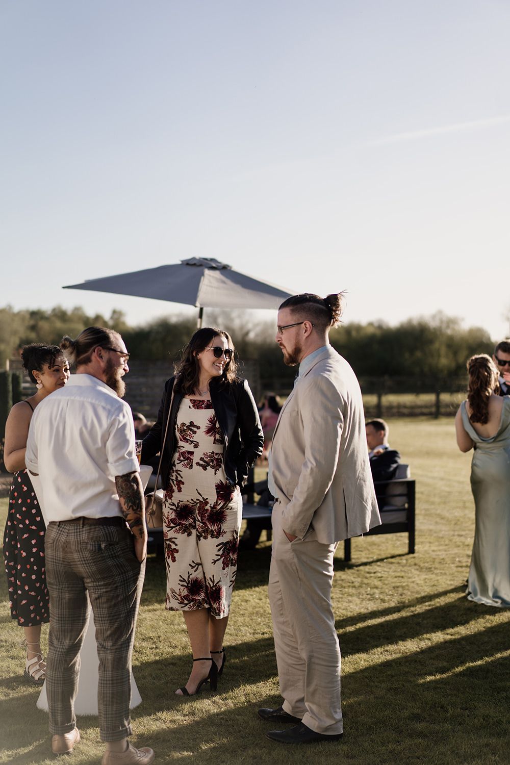 Wedding guests stood talking while standing on grass with the sun behind them.