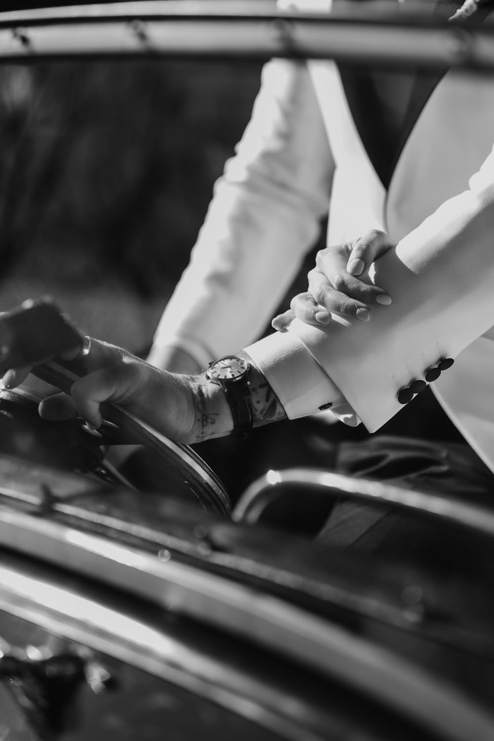Black and white photo of a groom helping bride out of a vintage car, showcasing a watch and tattoo on his wrist.