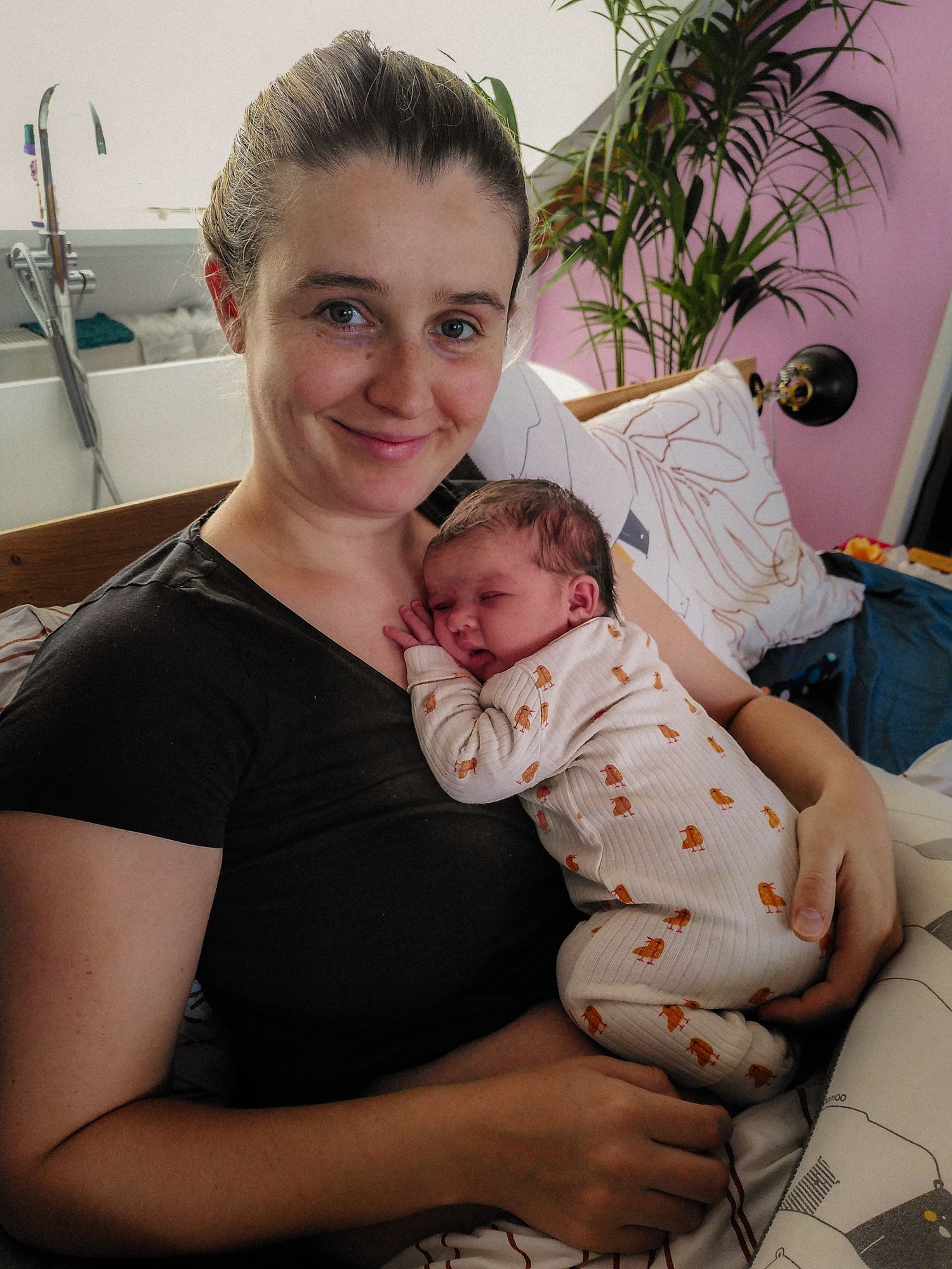 A smiling woman in a black shirt holds a sleeping newborn baby wrapped in a white outfit with small prints.
