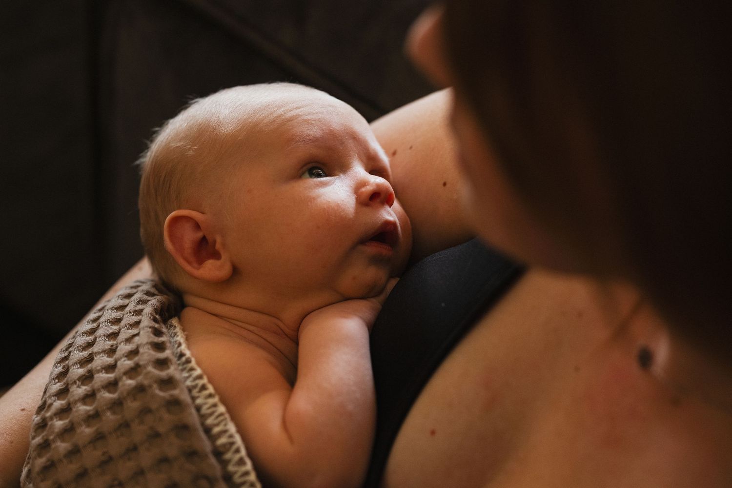 Newborn baby gazing up at mother during breastfeeding, wrapped in a soft blanket in warm, dim lighting.