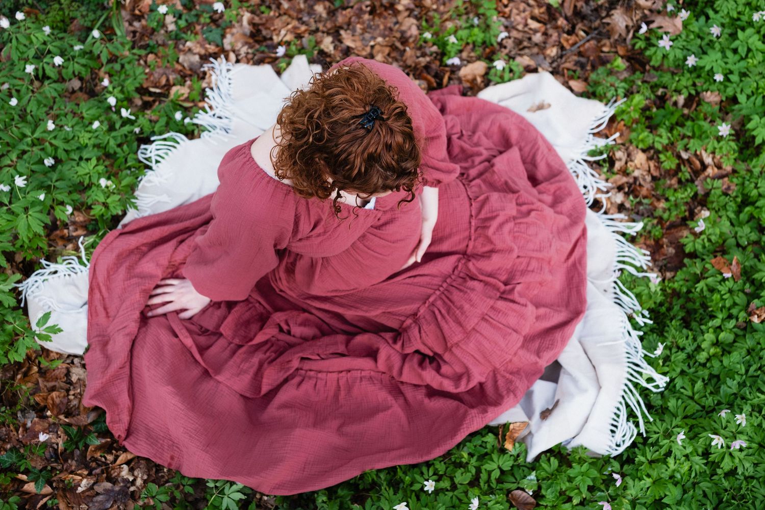 Woman in a flowing pink dress sitting on a white blanket surrounded by green grass and small white flowers.