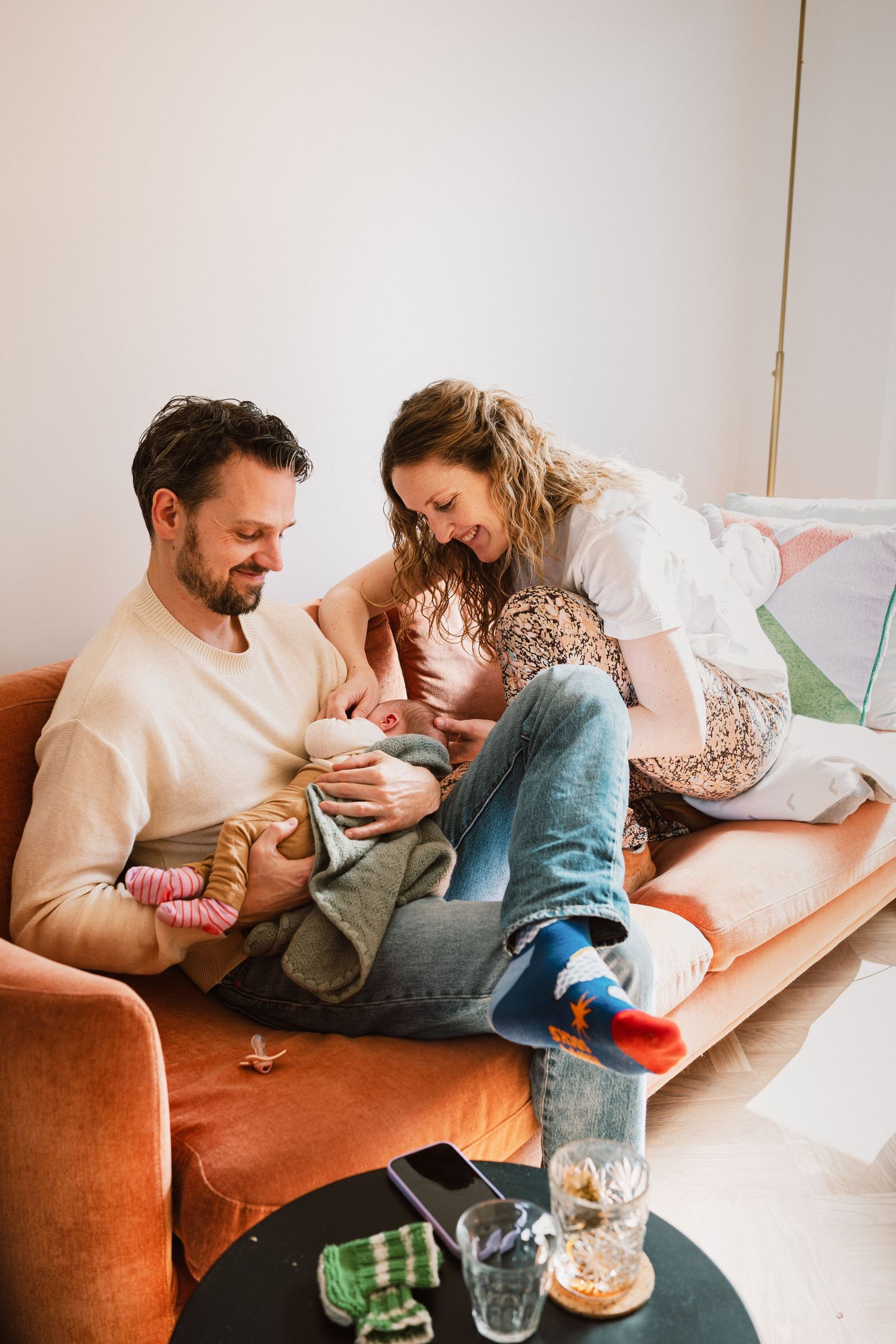 Happy couple sitting on orange sofa, lovingly cradling their newborn baby together in a bright living room.