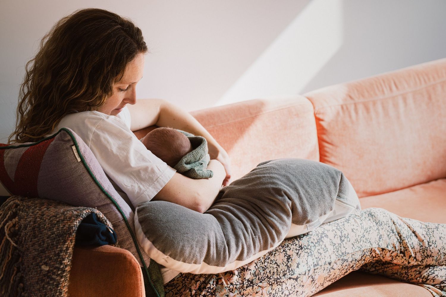 A mother sits on a pink sofa, lovingly cradling and breastfeeding her newborn baby wrapped in a blanket.