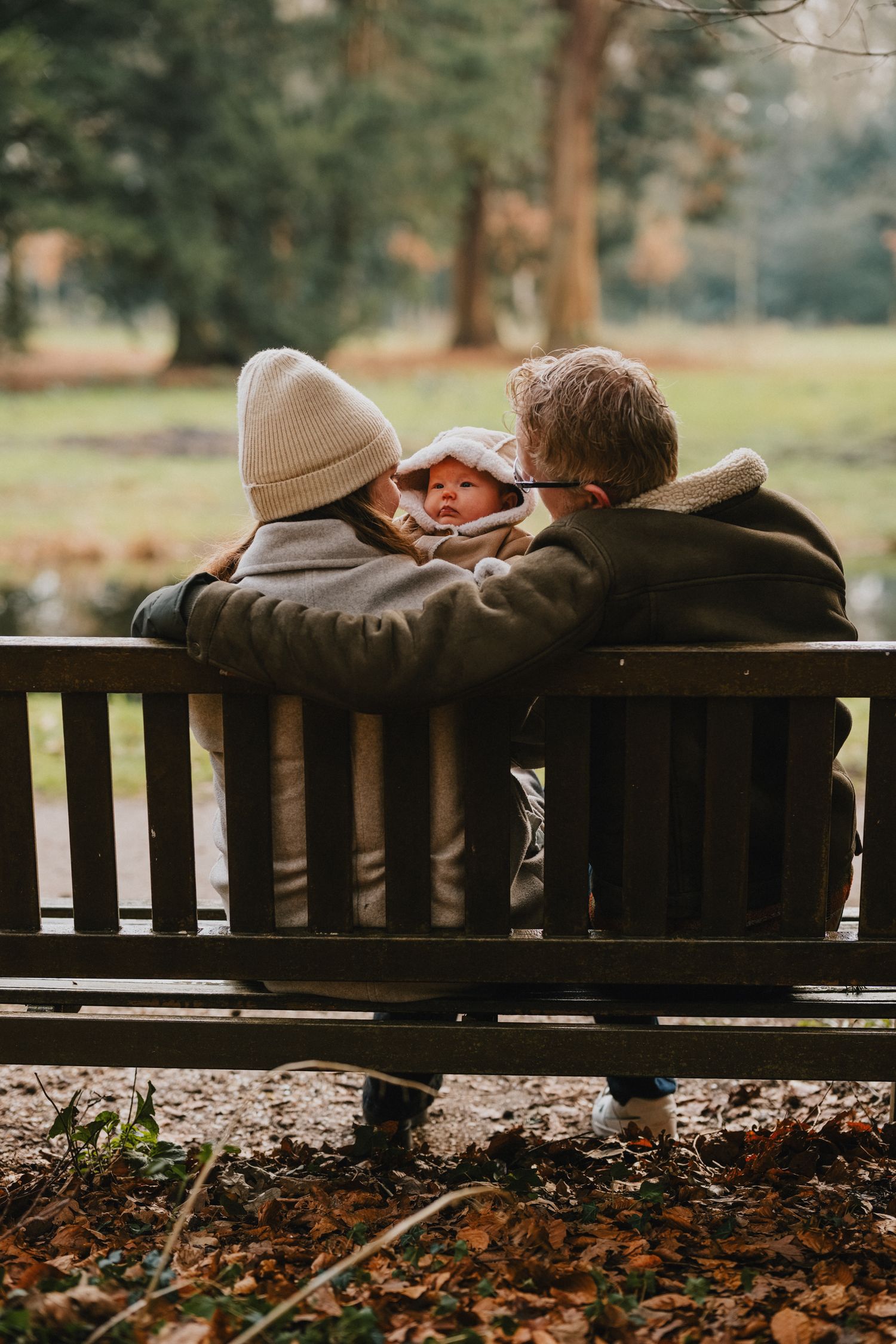 Two people sit close together on a wooden park bench surrounded by autumn leaves and tall pine trees in the background.