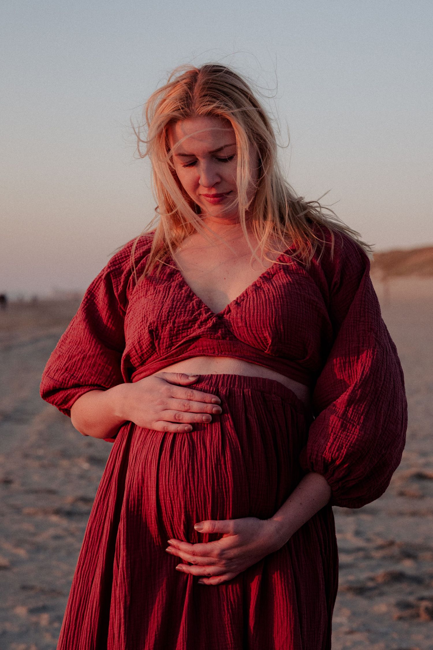 Pregnant woman in red dress cradling baby bump on beach at sunset with golden light.