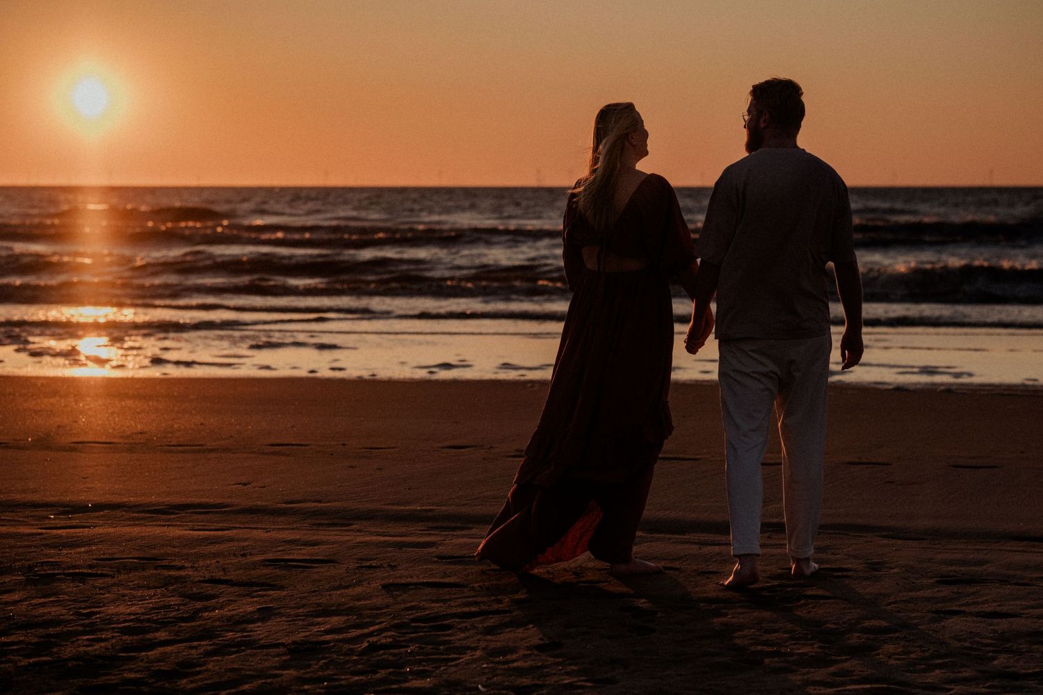Couple holding hands walking on a sandy beach at sunset with golden orange sky and ocean waves in background.