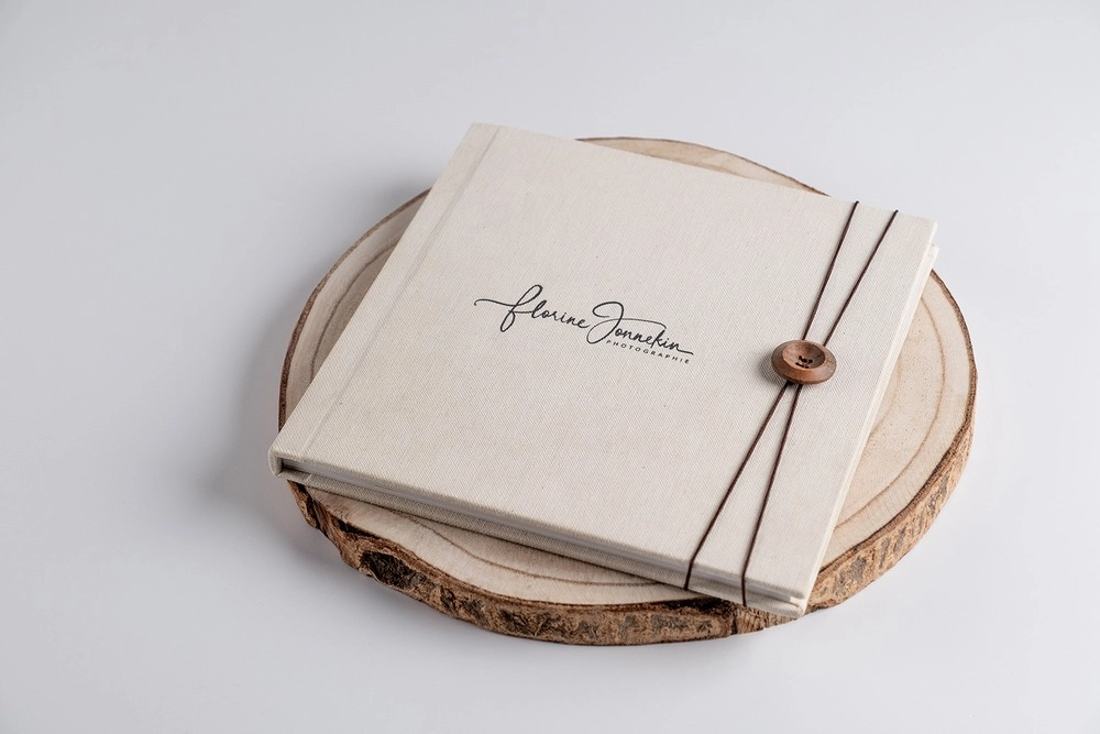 Elegant linen photo album with script logo, tied with brown cord and button, displayed on a rustic wood slice.