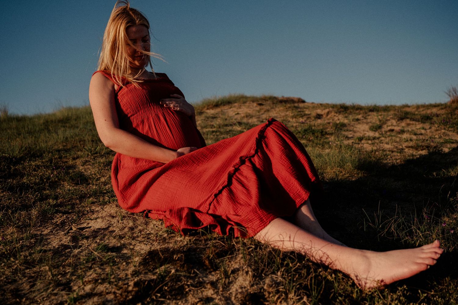 Pregnant woman in red dress sitting barefoot on grassy hillside in warm sunlight.