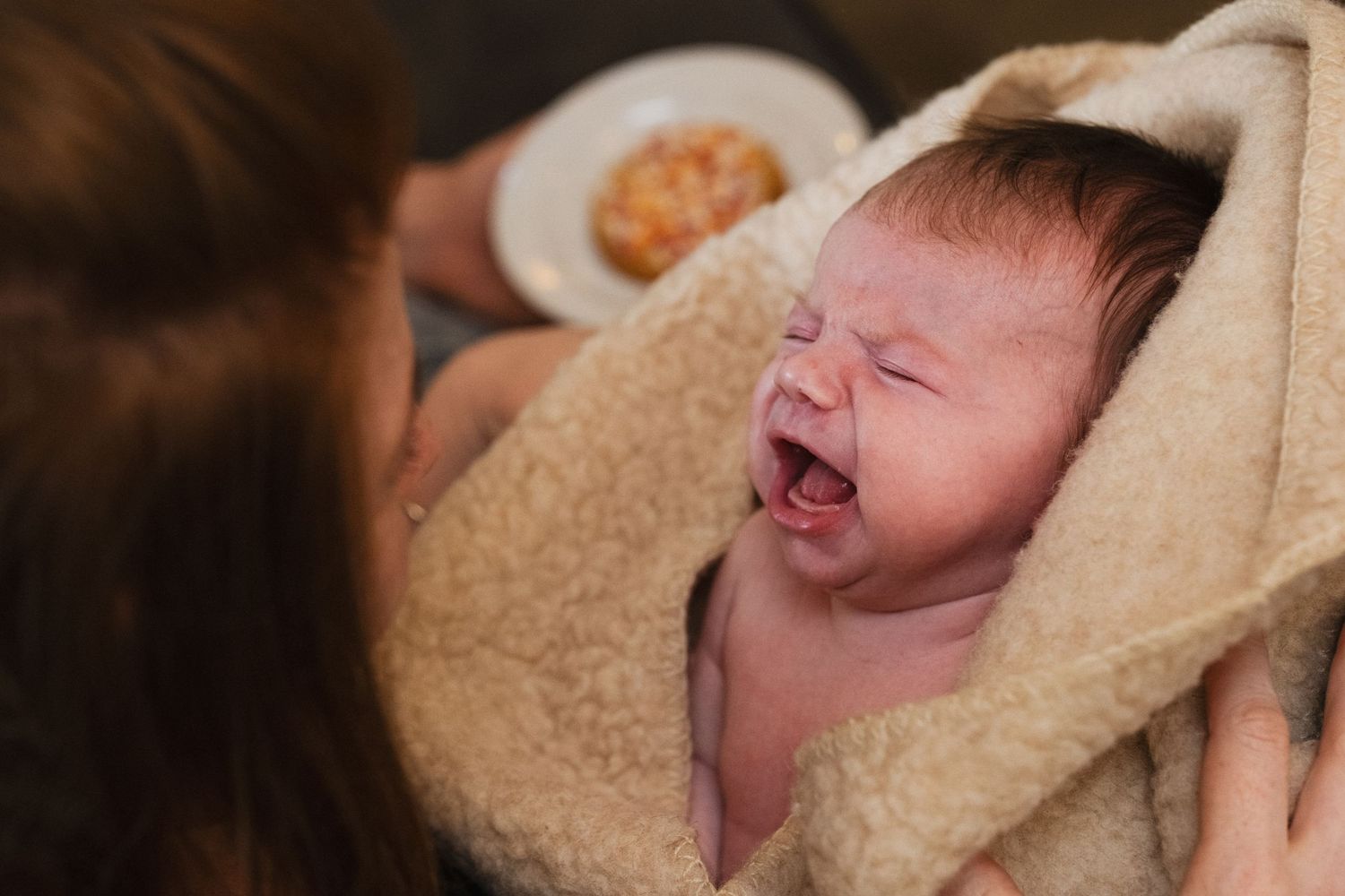 Newborn baby wrapped in a soft beige towel yawning after bath, held by mother.
