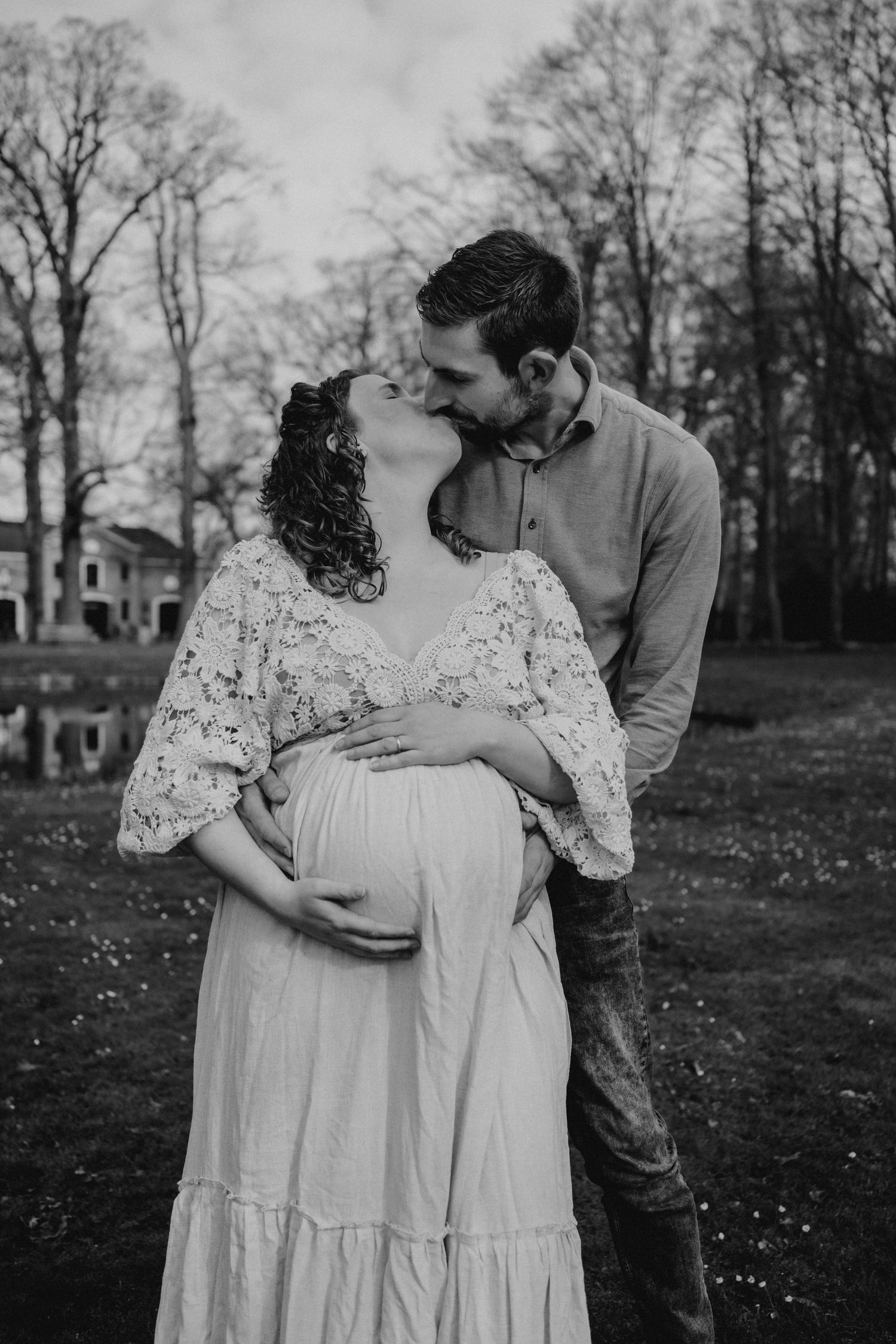 Black and white maternity photo of a couple embracing outdoors, man kissing pregnant woman in lace dress.