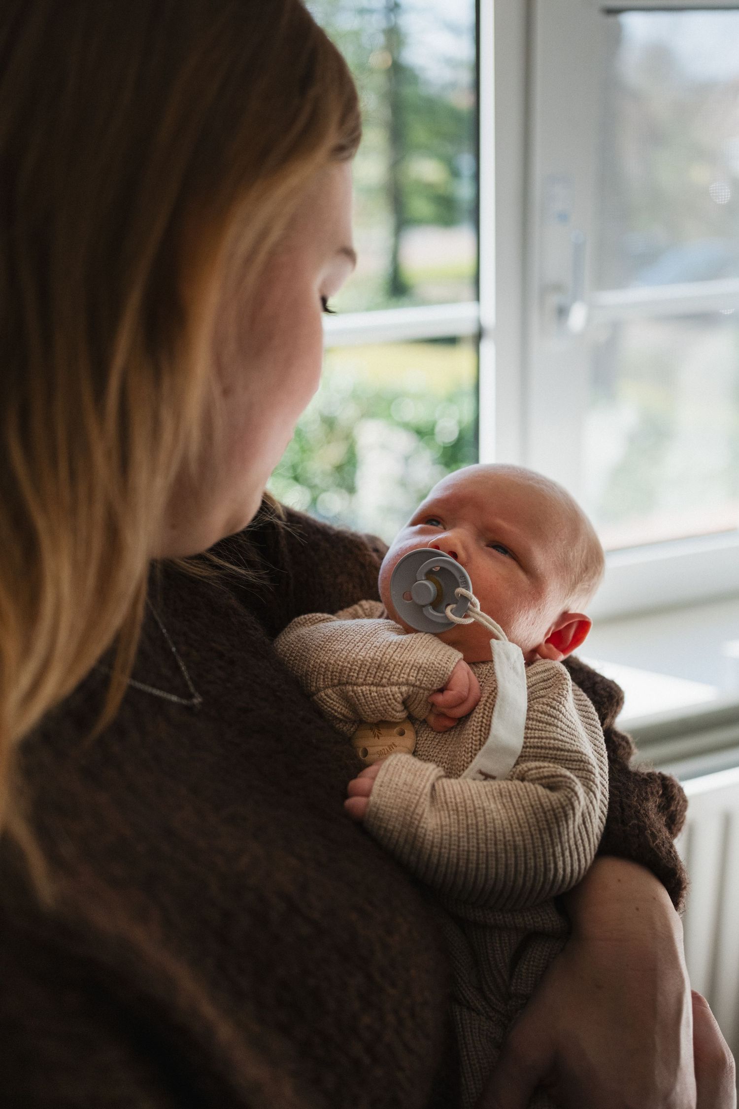 A mother holds her newborn baby with a pacifier near a bright window, wrapped in a cozy beige knit outfit.