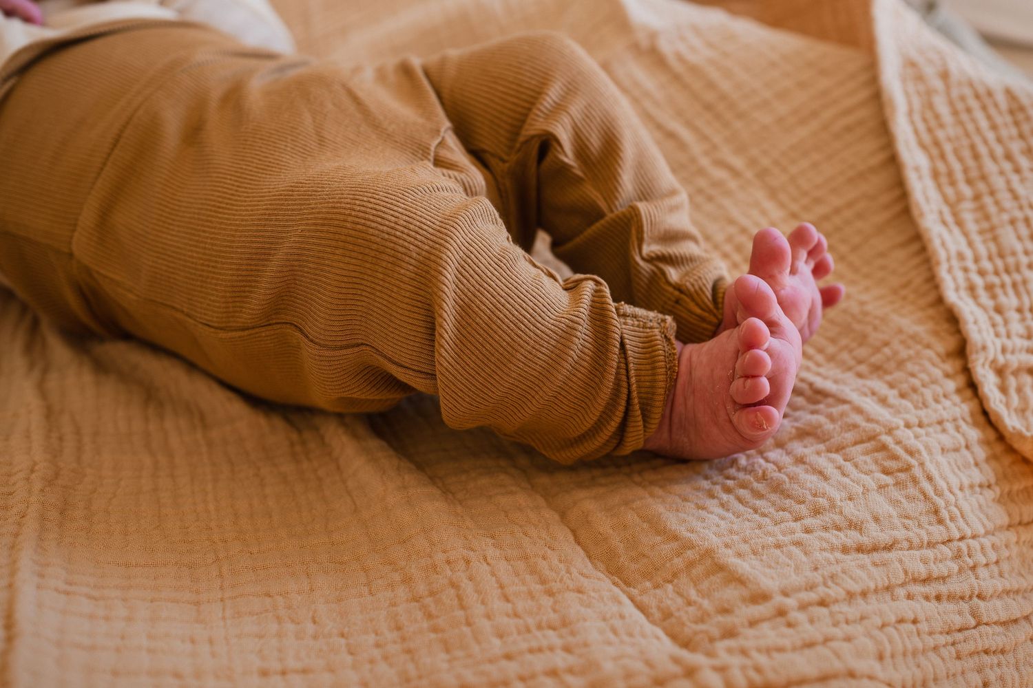 Close-up of a newborn baby's tiny bare feet and brown ribbed pants lying on a soft peach muslin blanket.