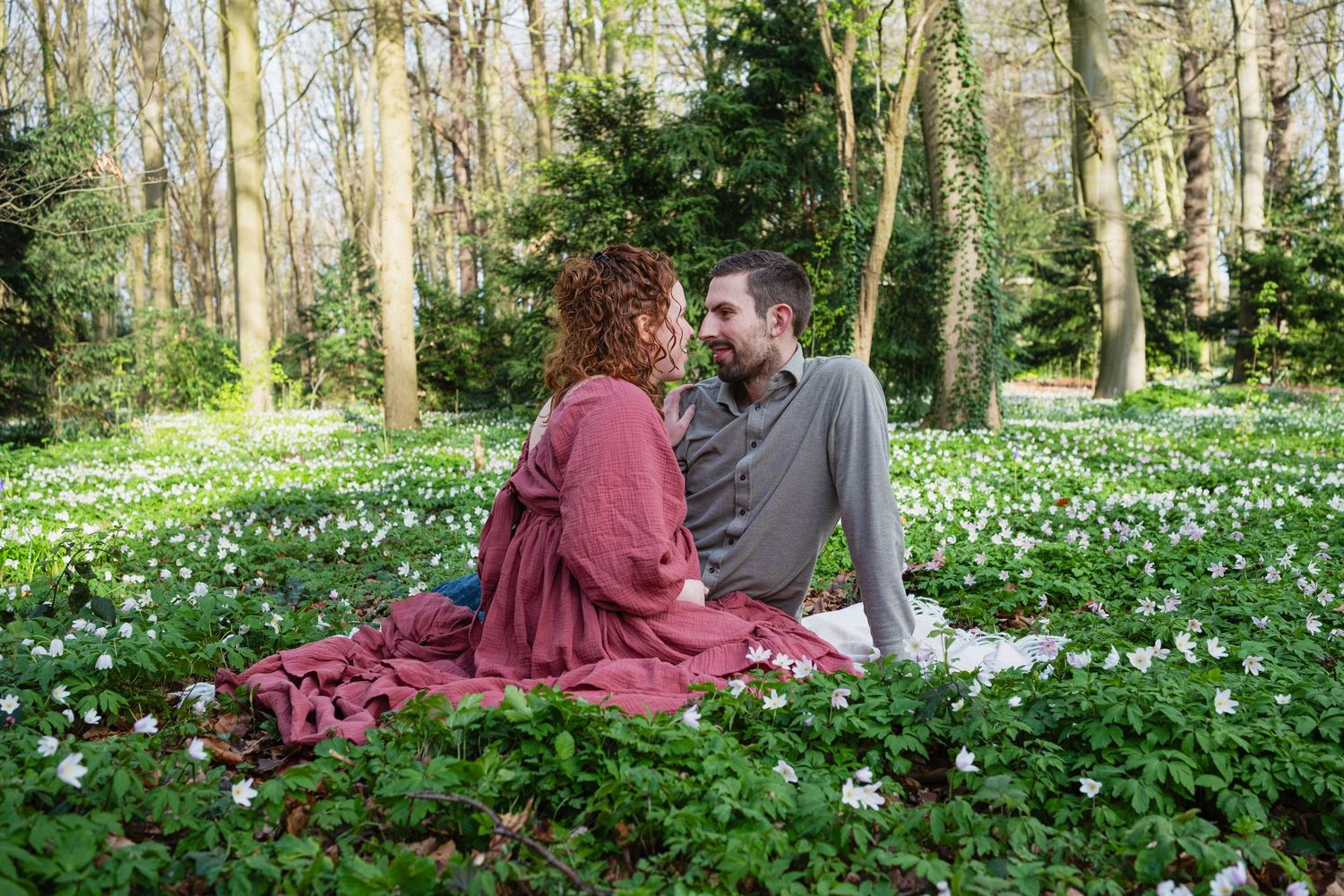 Couple sitting together in a forest meadow filled with white flowers on a red blanket in spring.