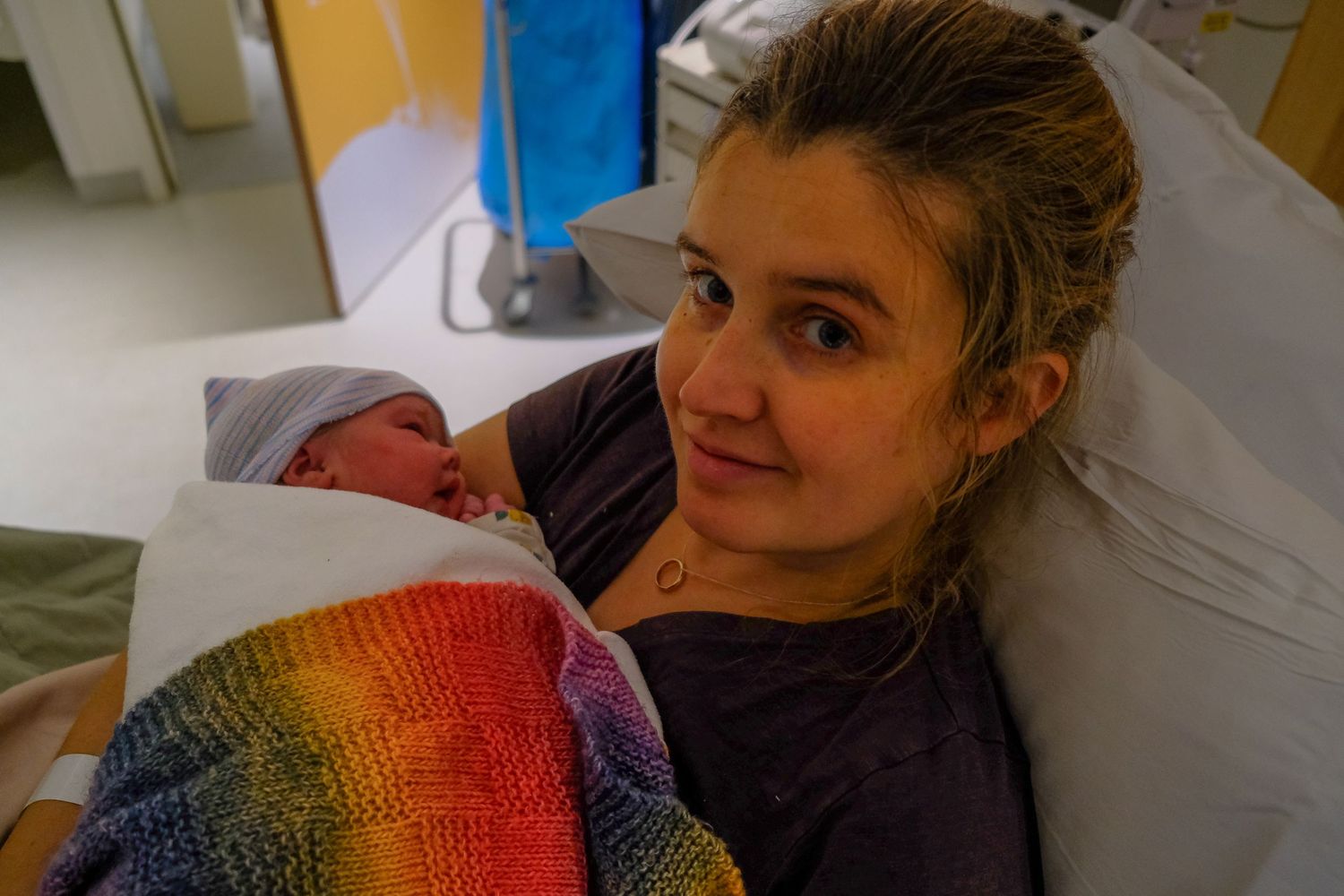 New mother smiling in hospital bed holding newborn baby wrapped in colorful rainbow knit blanket.