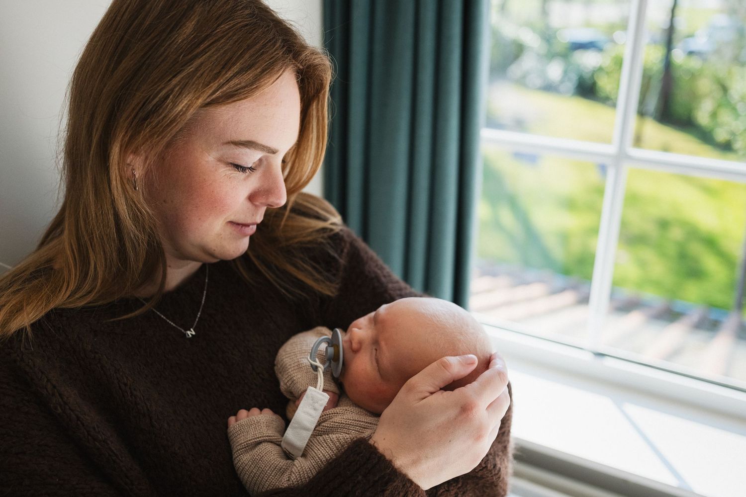 A young mother tenderly holds her newborn baby near a window in a warm, intimate portrait.