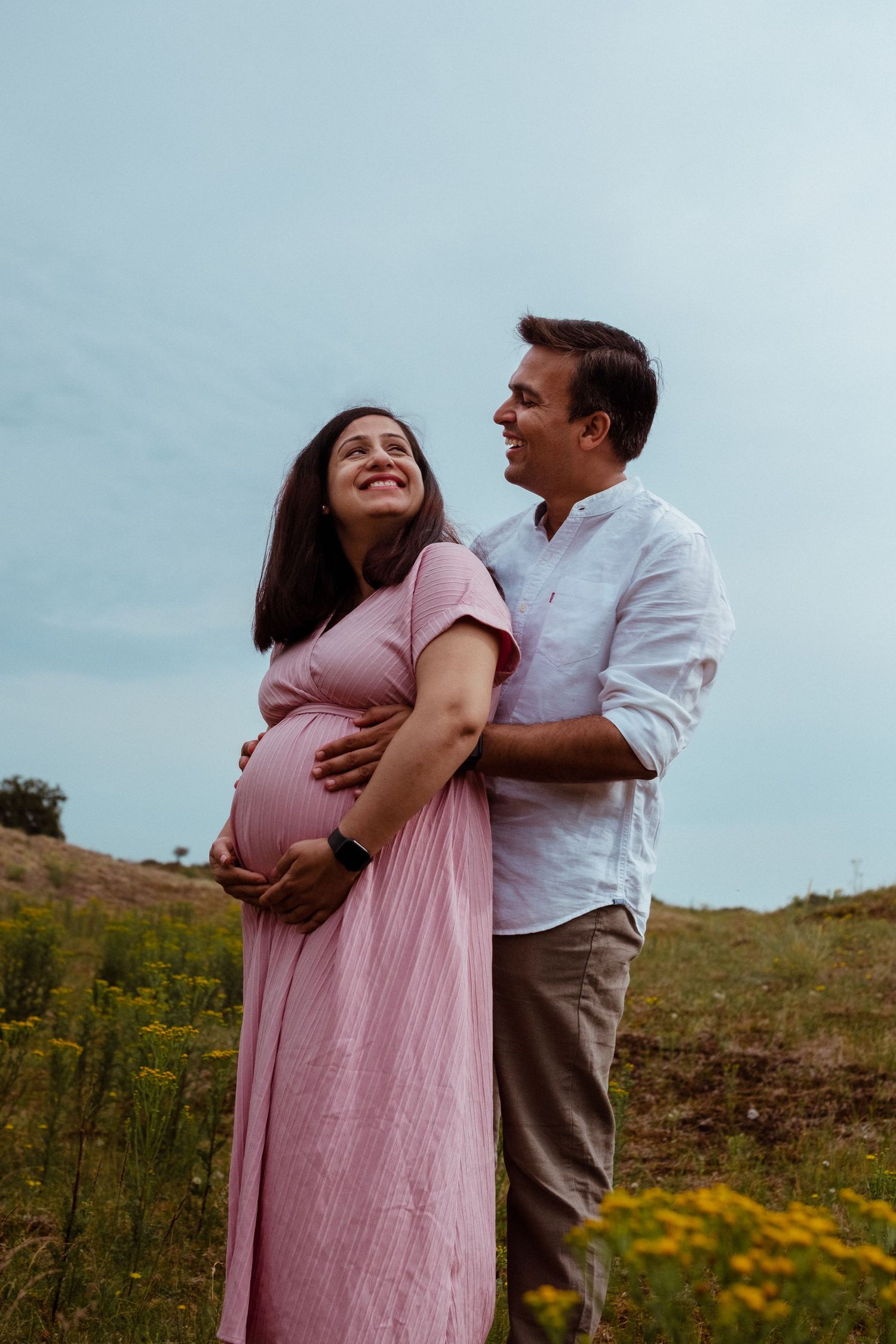 Smiling pregnant woman in pink dress embraced by man in white shirt standing in a green field.