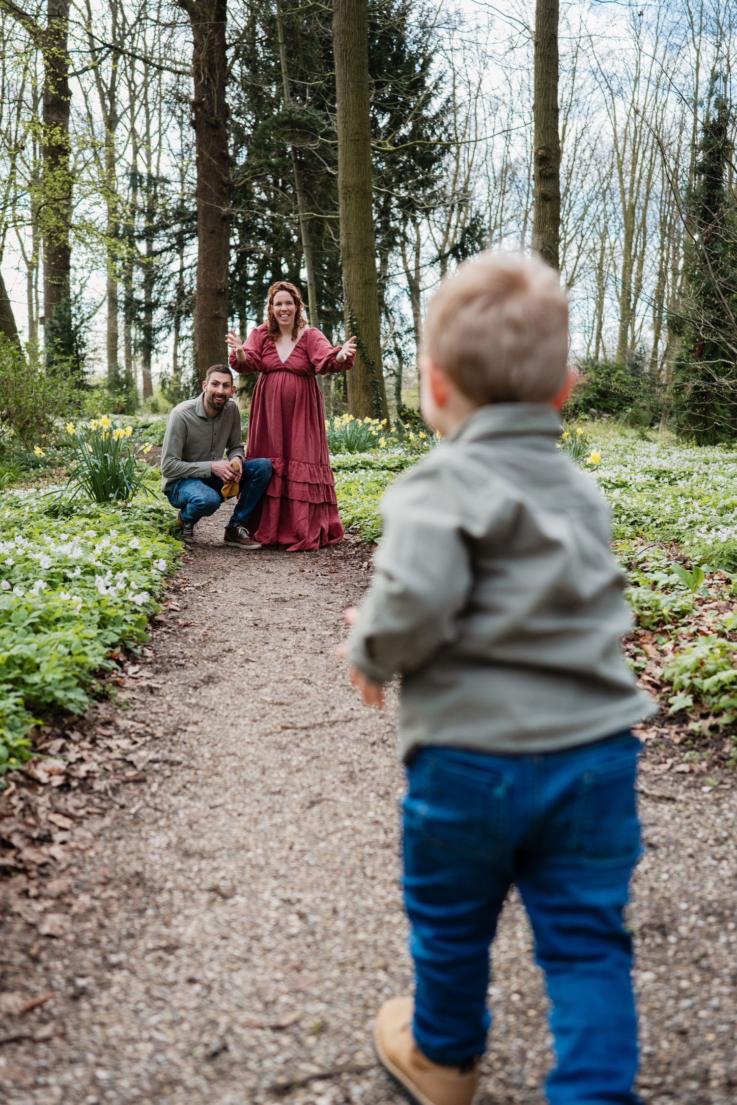 A young child walks toward parents on a forest path, mother in red dress with arms open, surrounded by spring flowers.